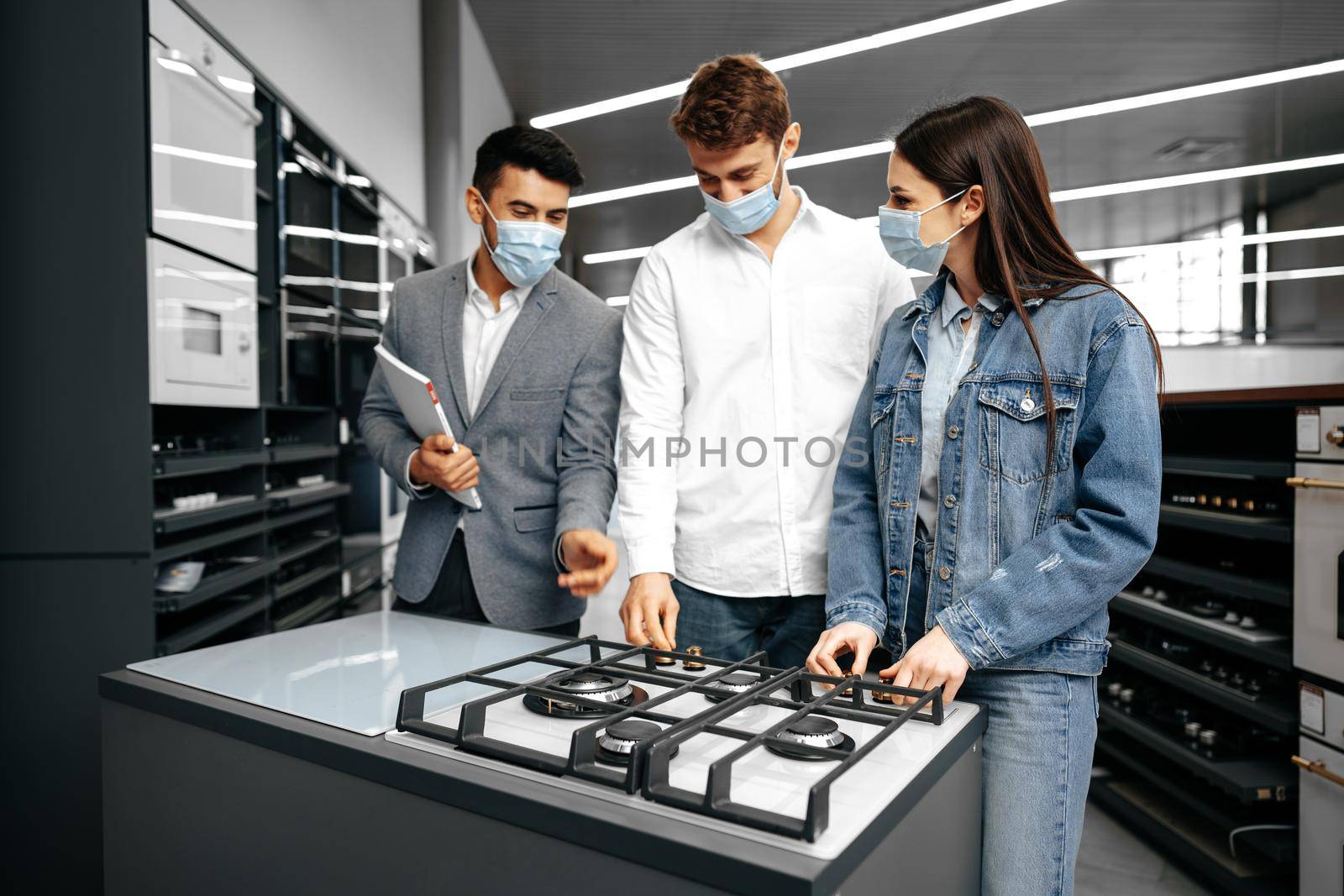 Shop assistant in hypermarket shows new model of gas stove to young couple, all wearing medical masks, close up