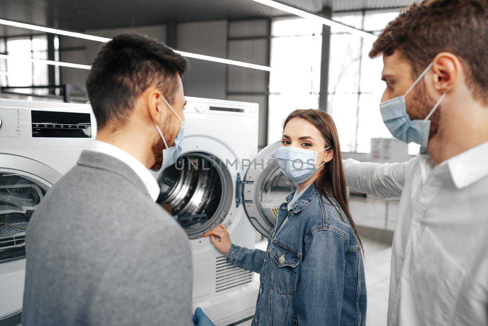 Salesman in hypermarket wearing medical mask demonstrates his clients a new washing machine by Fabrikasimf
