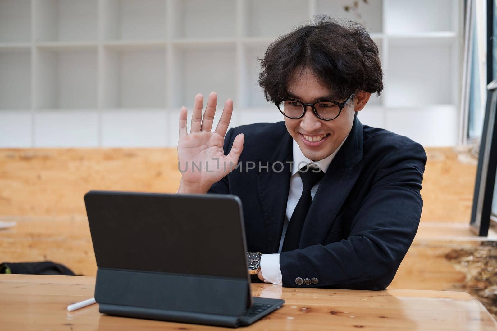 A young asian business man working remotely heaving a meeting on a digital tablet. man waving hand at home during self isolation from coronavirus outbreak crisis.
