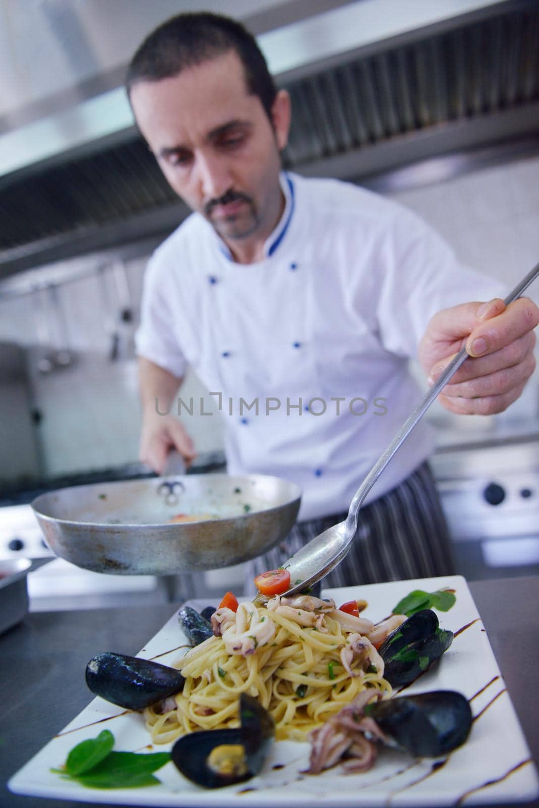 Handsome chef dressed in white uniform decorating pasta salad and seafood fish in modern kitchen