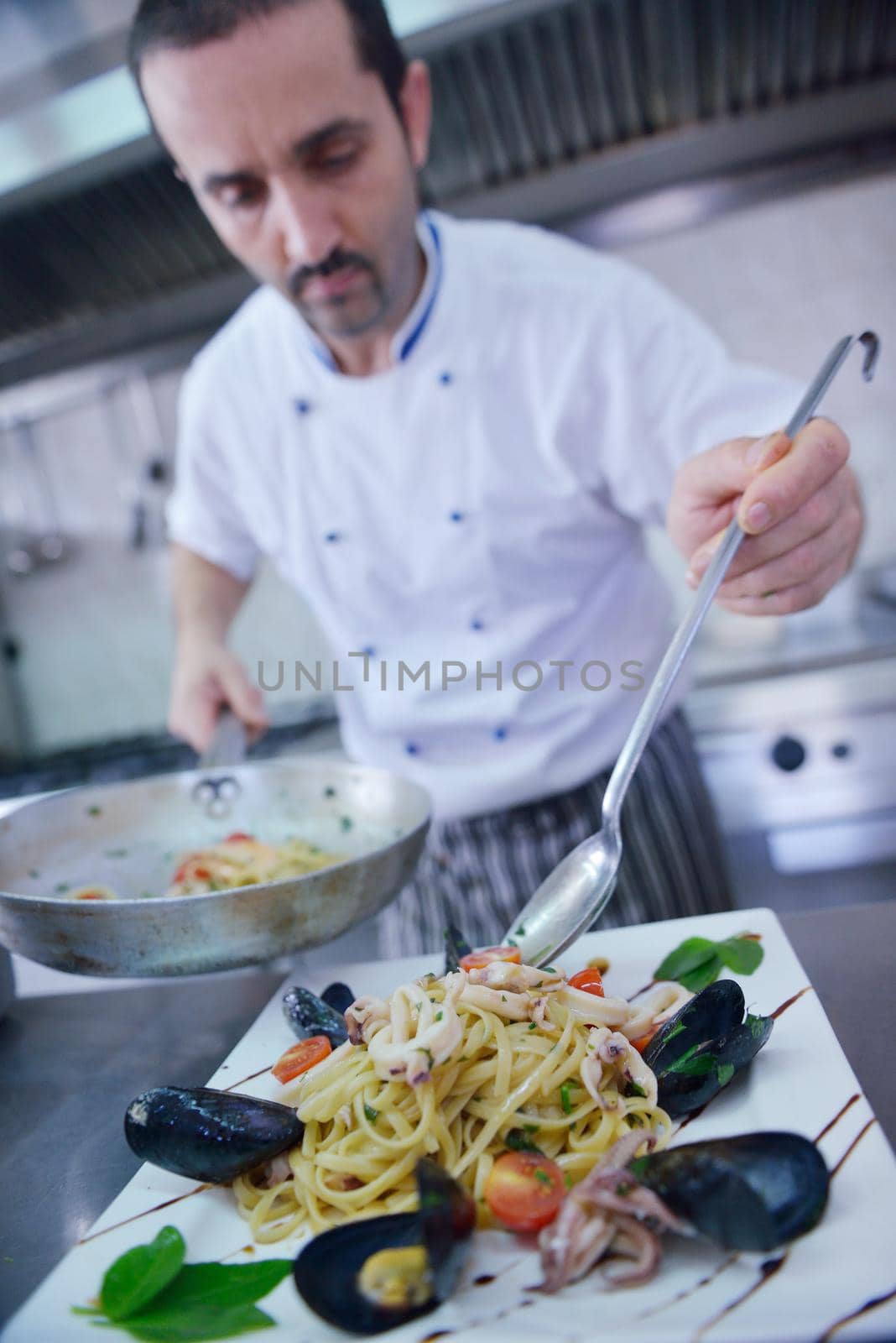 Handsome chef dressed in white uniform decorating pasta salad and seafood fish in modern kitchen