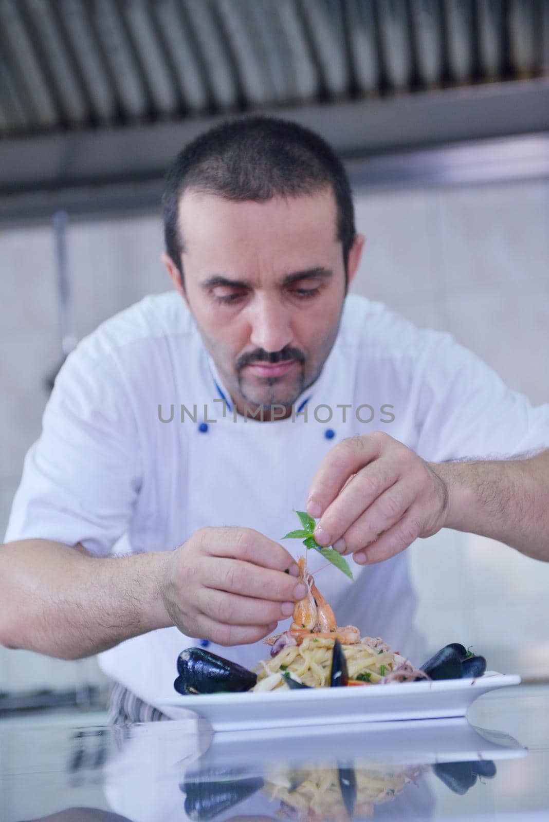 Handsome chef dressed in white uniform decorating pasta salad and seafood fish in modern kitchen