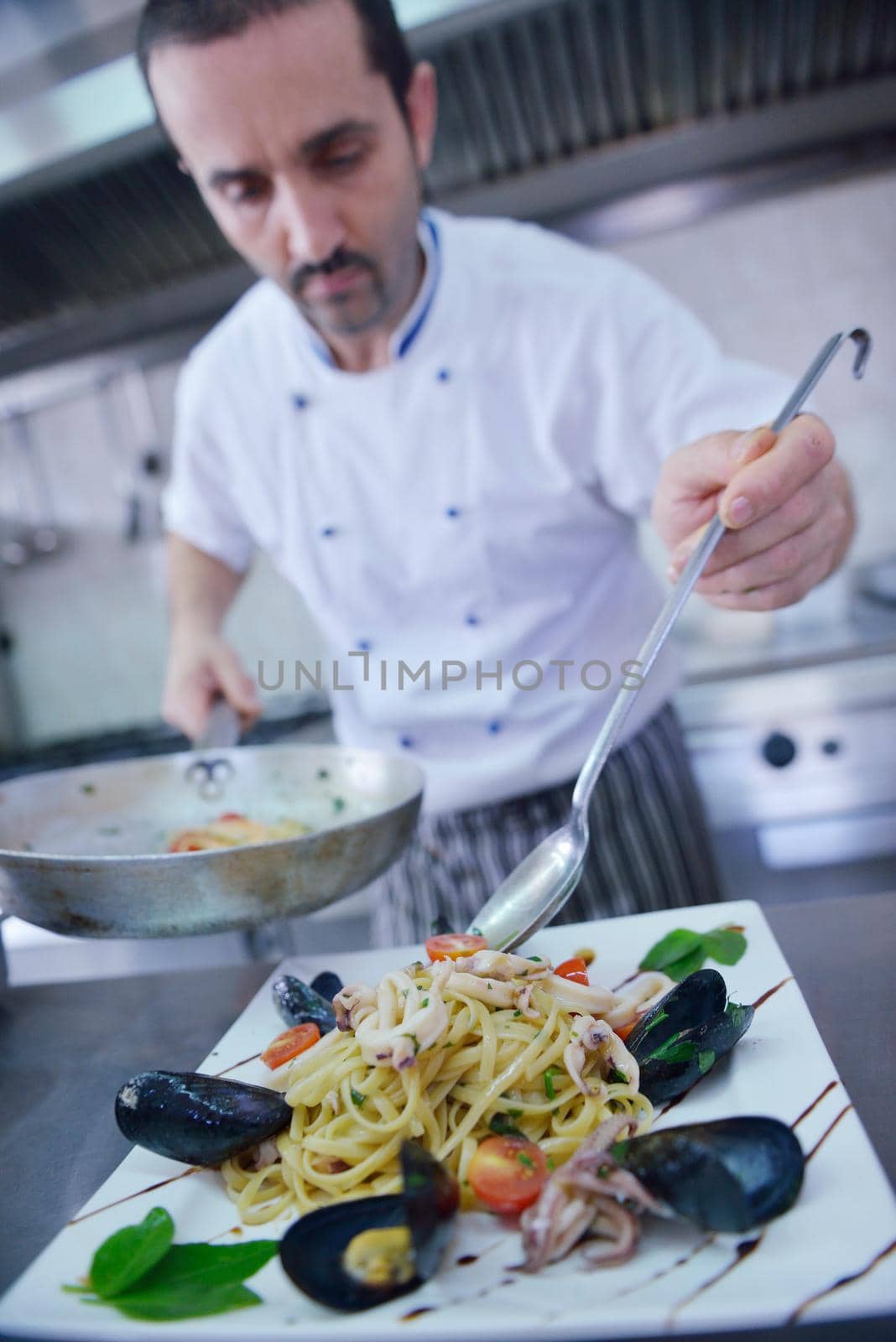 Handsome chef dressed in white uniform decorating pasta salad and seafood fish in modern kitchen