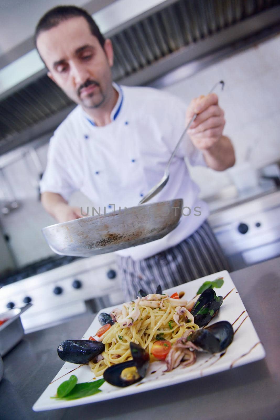 Handsome chef dressed in white uniform decorating pasta salad and seafood fish in modern kitchen