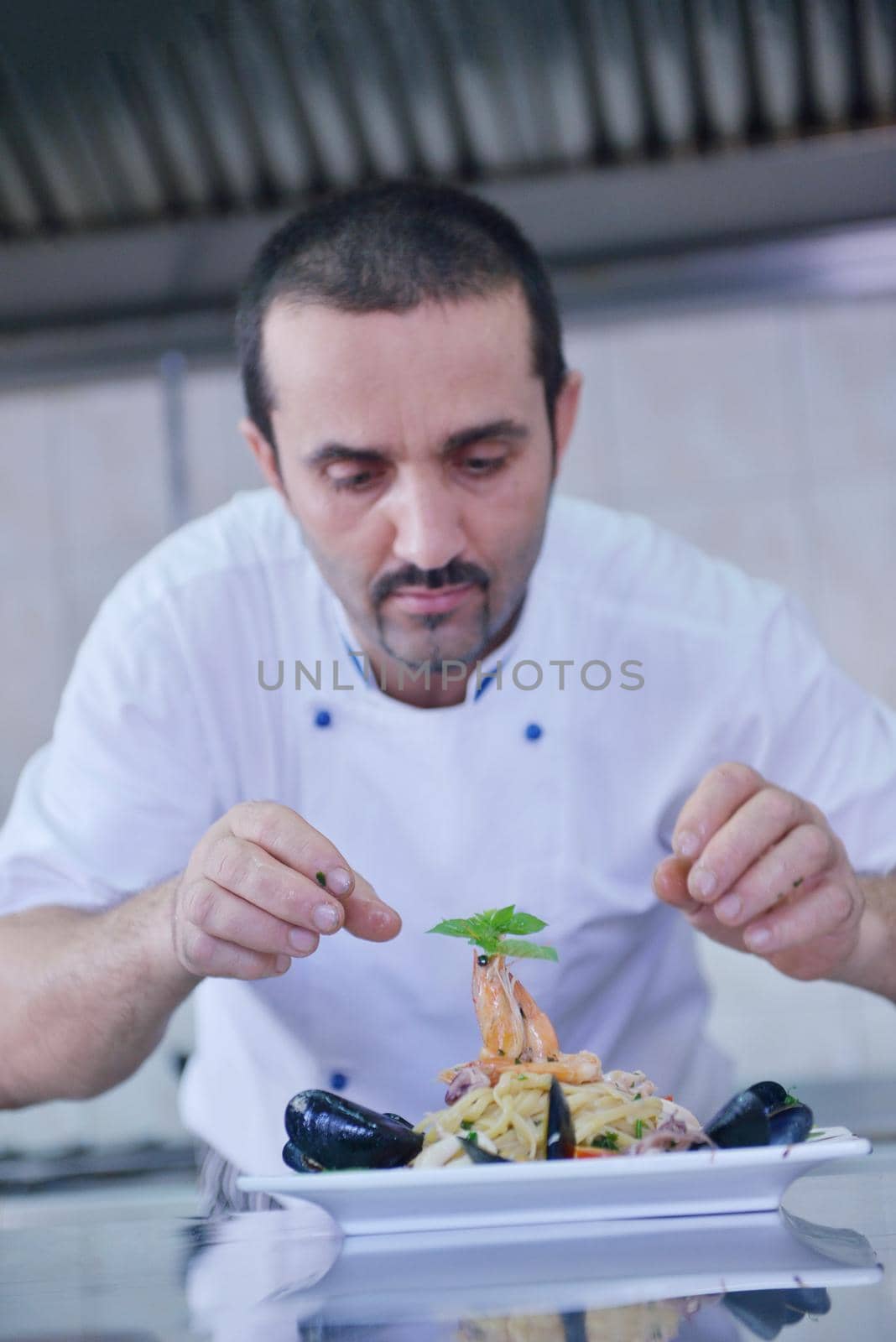 Handsome chef dressed in white uniform decorating pasta salad and seafood fish in modern kitchen