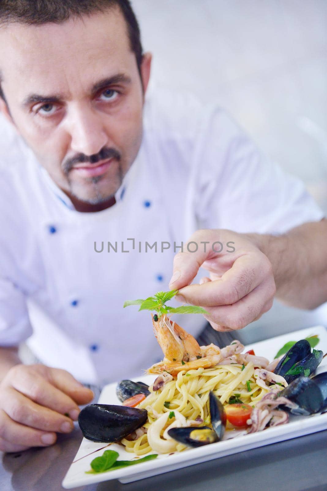 Handsome chef dressed in white uniform decorating pasta salad and seafood fish in modern kitchen