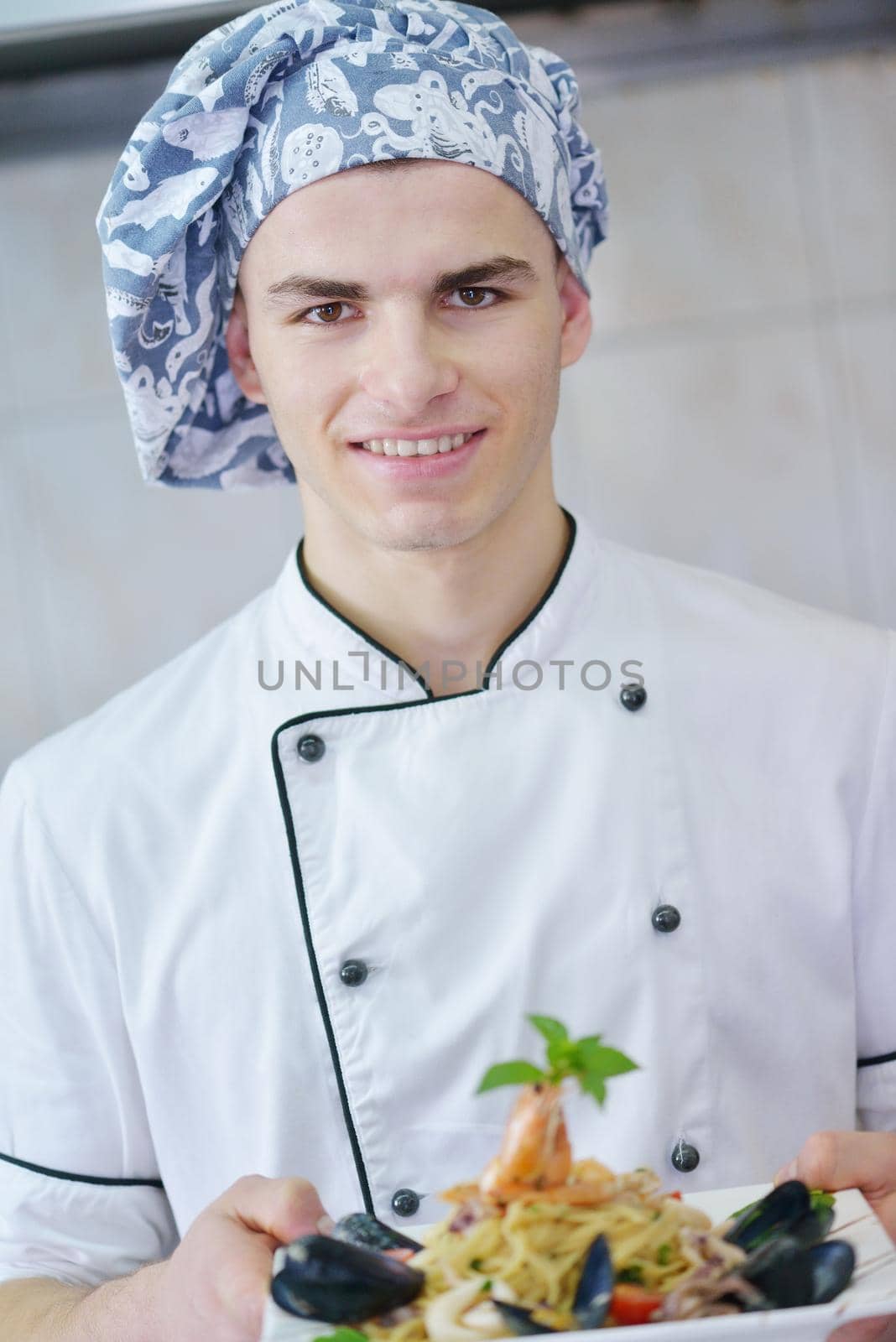 Handsome chef dressed in white uniform decorating pasta salad and seafood fish in modern kitchen