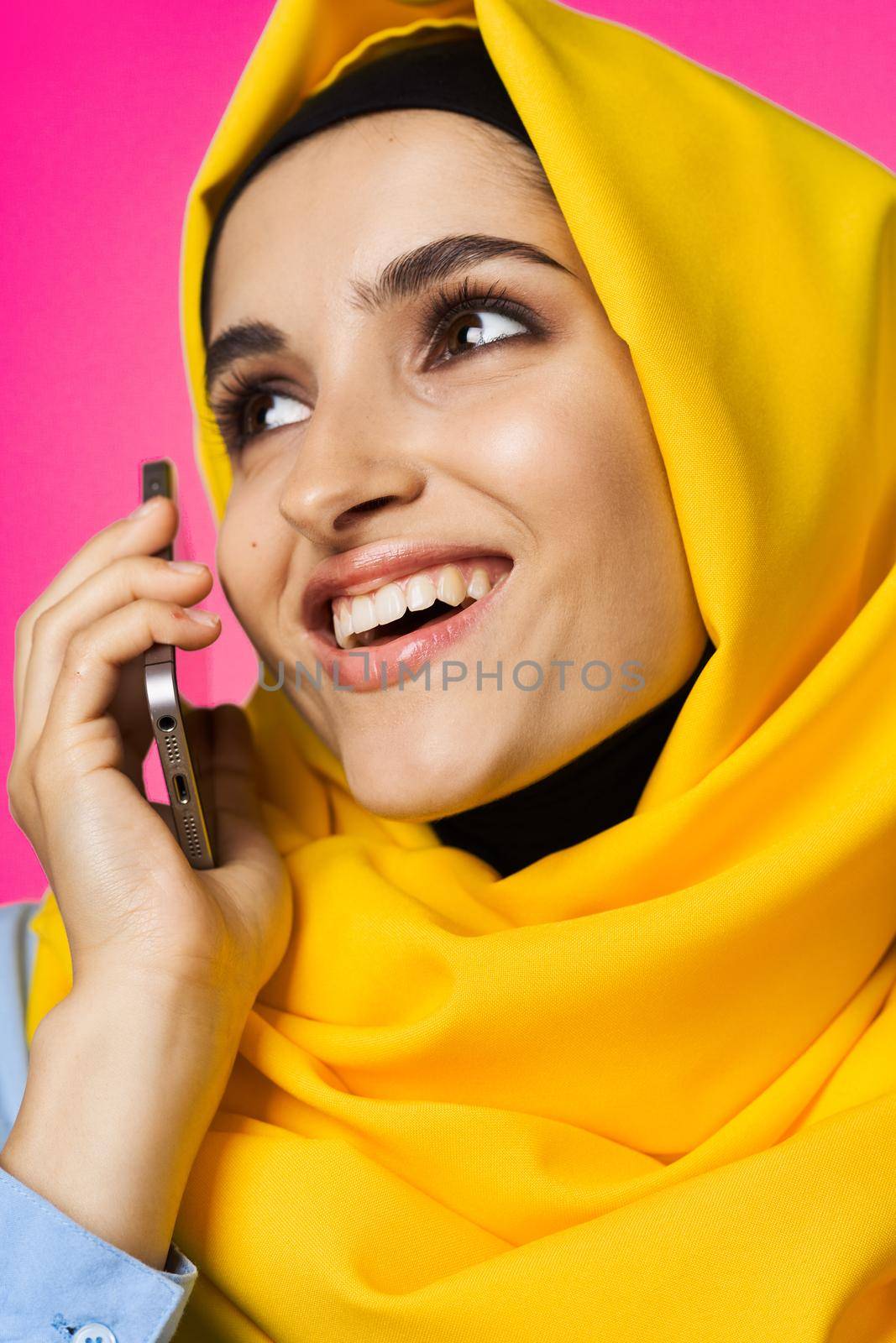 muslim woman in yellow hijab talking on the phone technology pink background. High quality photo