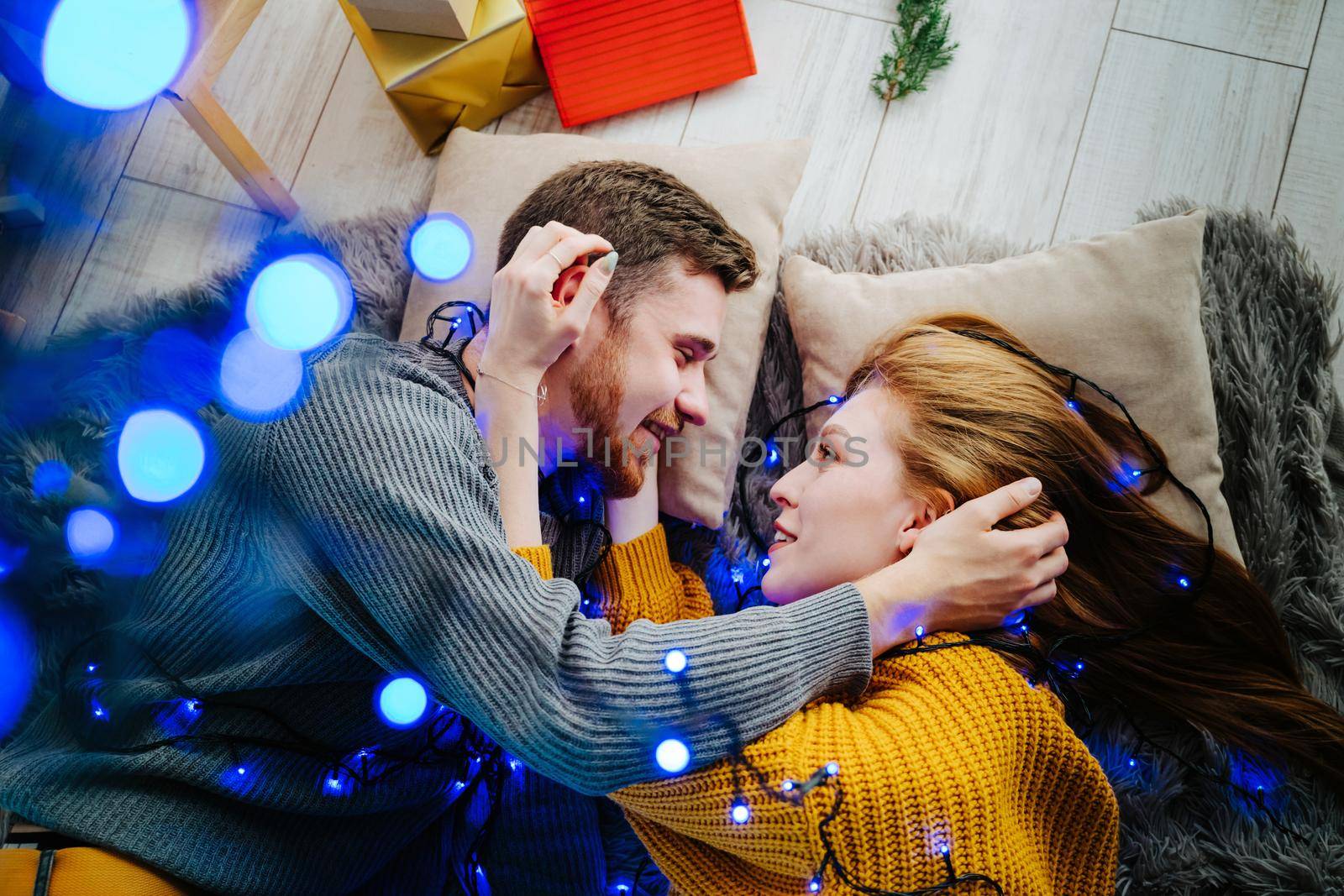 Beautiful couple in love lies on the floor and hugs. Blue bokeh. A happy guy and a girl lie on a soft gray blanket. Christmas blue garland.