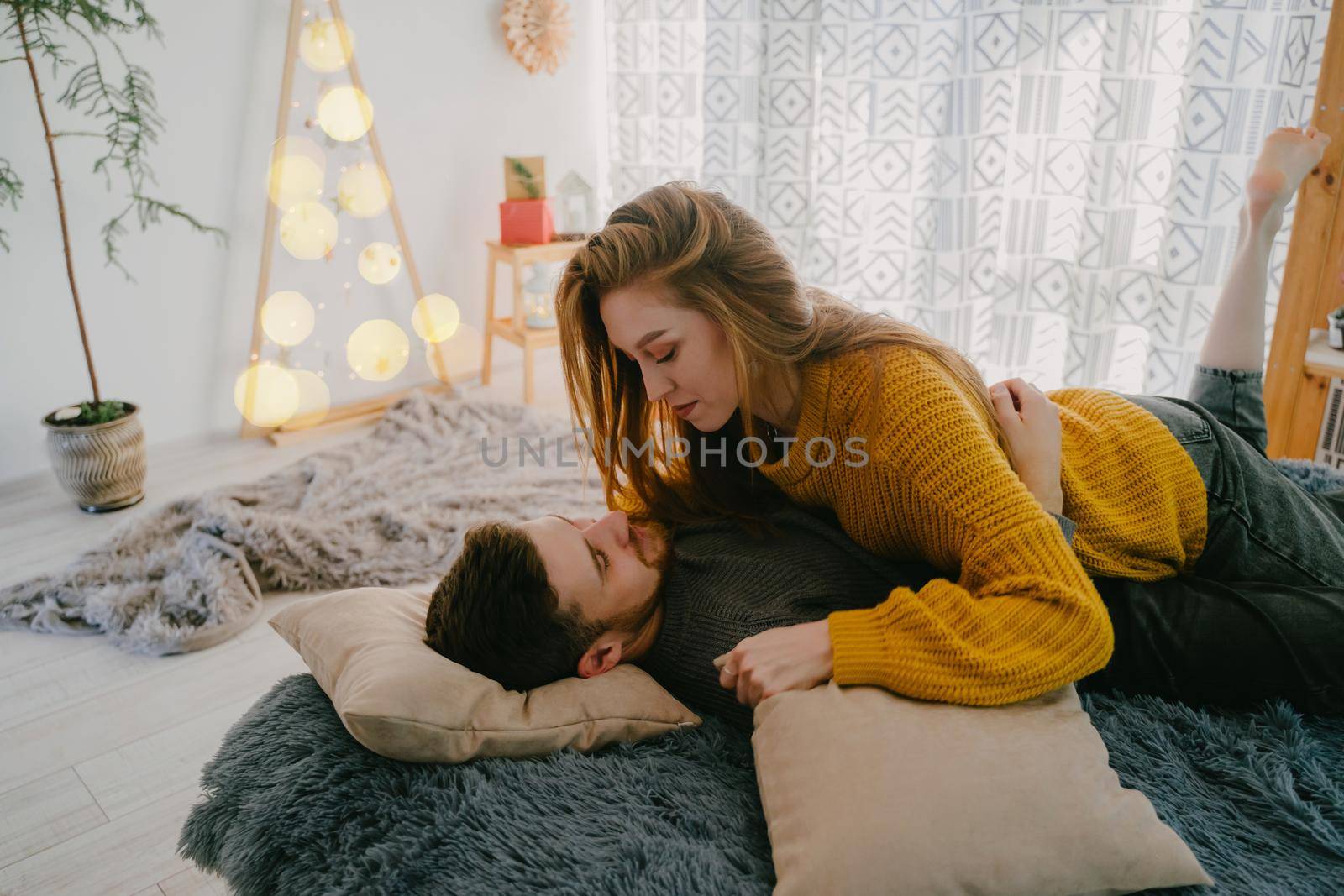 Beautiful couple in love lies on the bed against the background of Christmas decor. A live Christmas tree in a pot and a handmade Christmas tree made of natural wood. Christmas yellow garland.