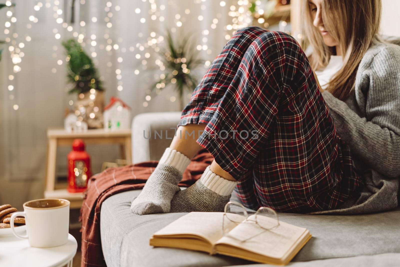 Cozy woman legs in knitted winter warm socks, sweater and checkered plaid drinking hot cocoa or coffee in mug, reading book, during resting on couch at home. Christmas holidays with decor and lights