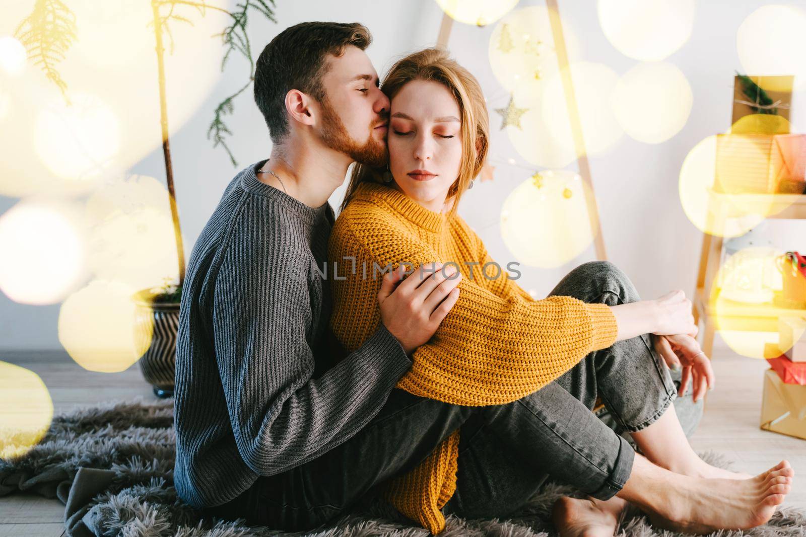 The guy gently kisses and hugs his girlfriend. Romantic couple of lovers against the background of a Christmas tree made of natural wood. Yellow bokeh.