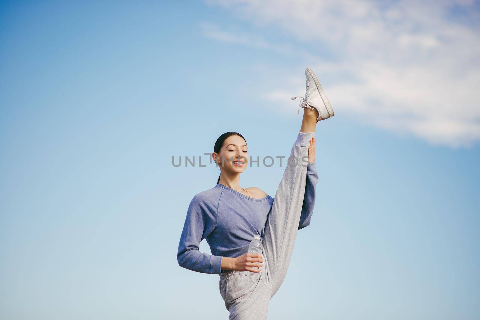 Beautiful girl training on a sky background. Woman in a blue sweater. Lady make a yoga. Girl with a bootle of water