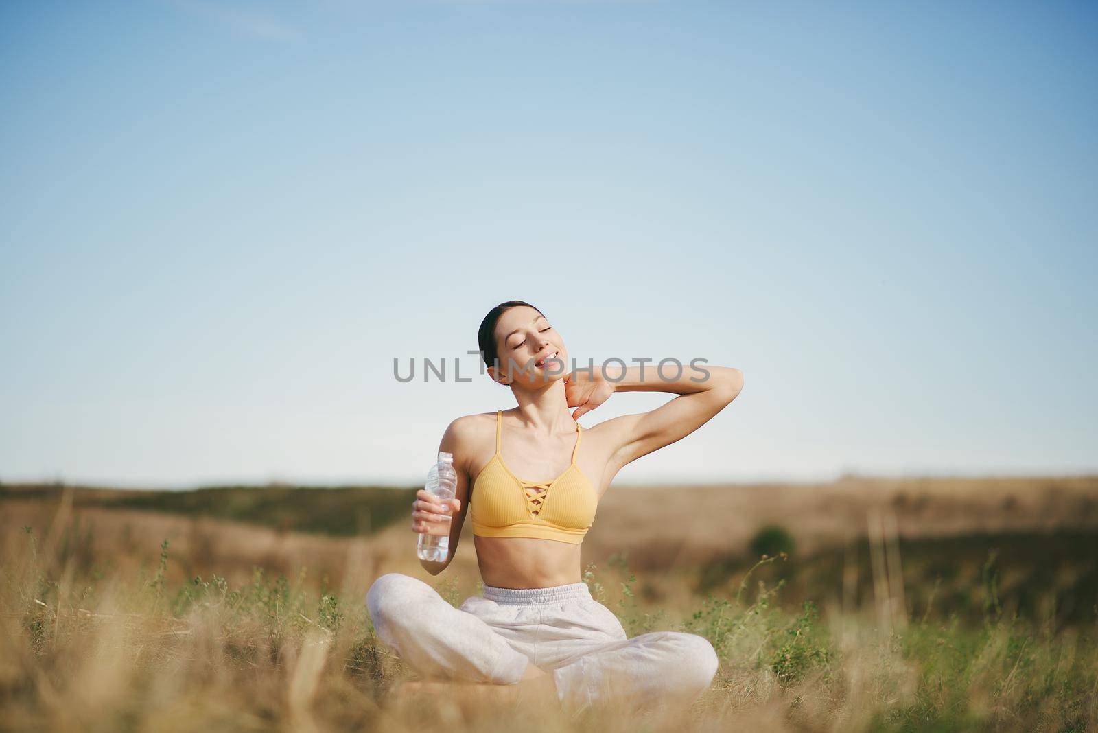 Beautiful girl training on a sky background. Woman in a yellow top. Lady make a yoga. Girl with a bootle of water.