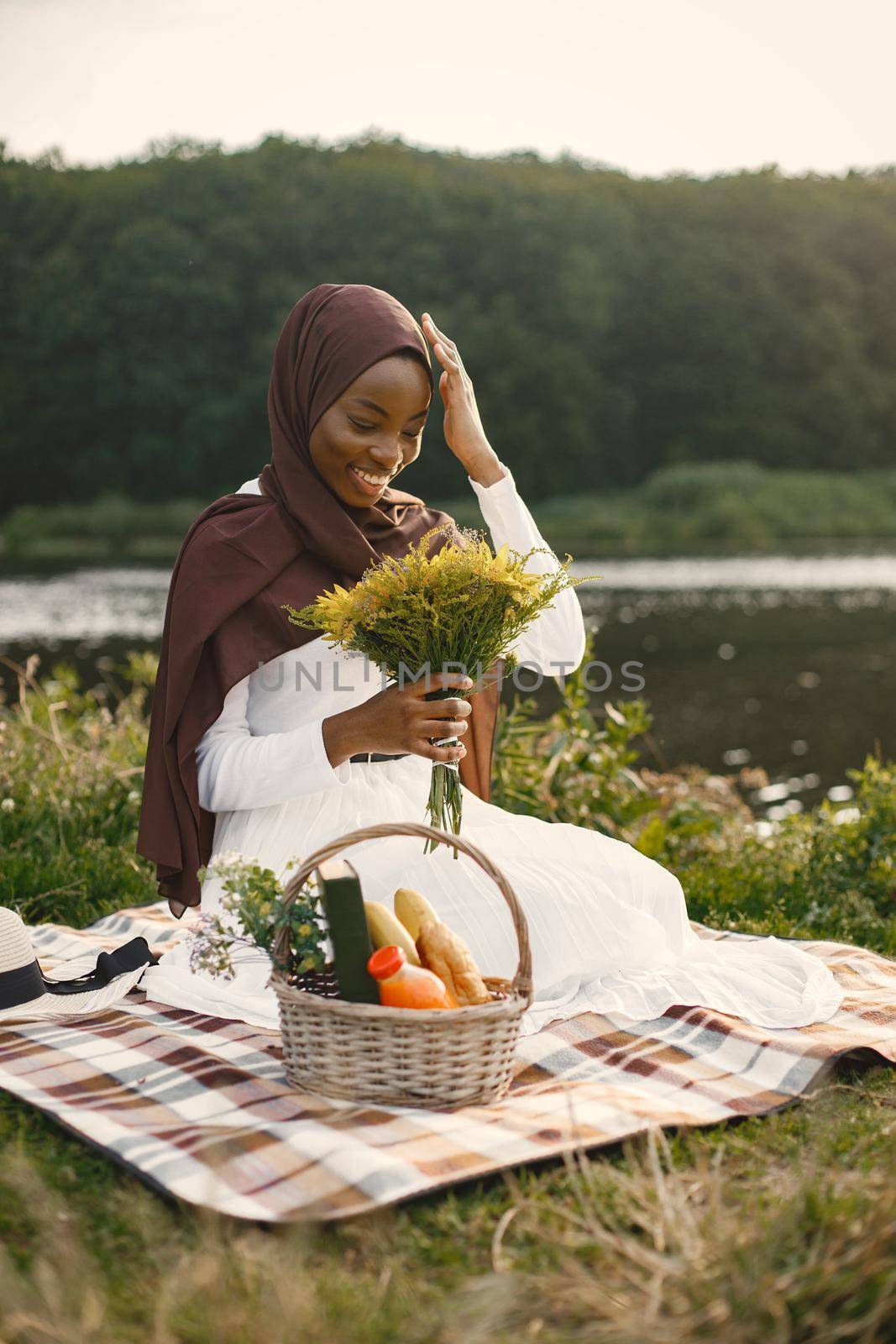 Muslim girl in hijab sits next to a basket with a picnic food holding a bouquet of flowers. Portrait of a Muslim lady in a brown hijab on the plaid picnic blanket near the river. Girl wearing white clothes and looking at camera.