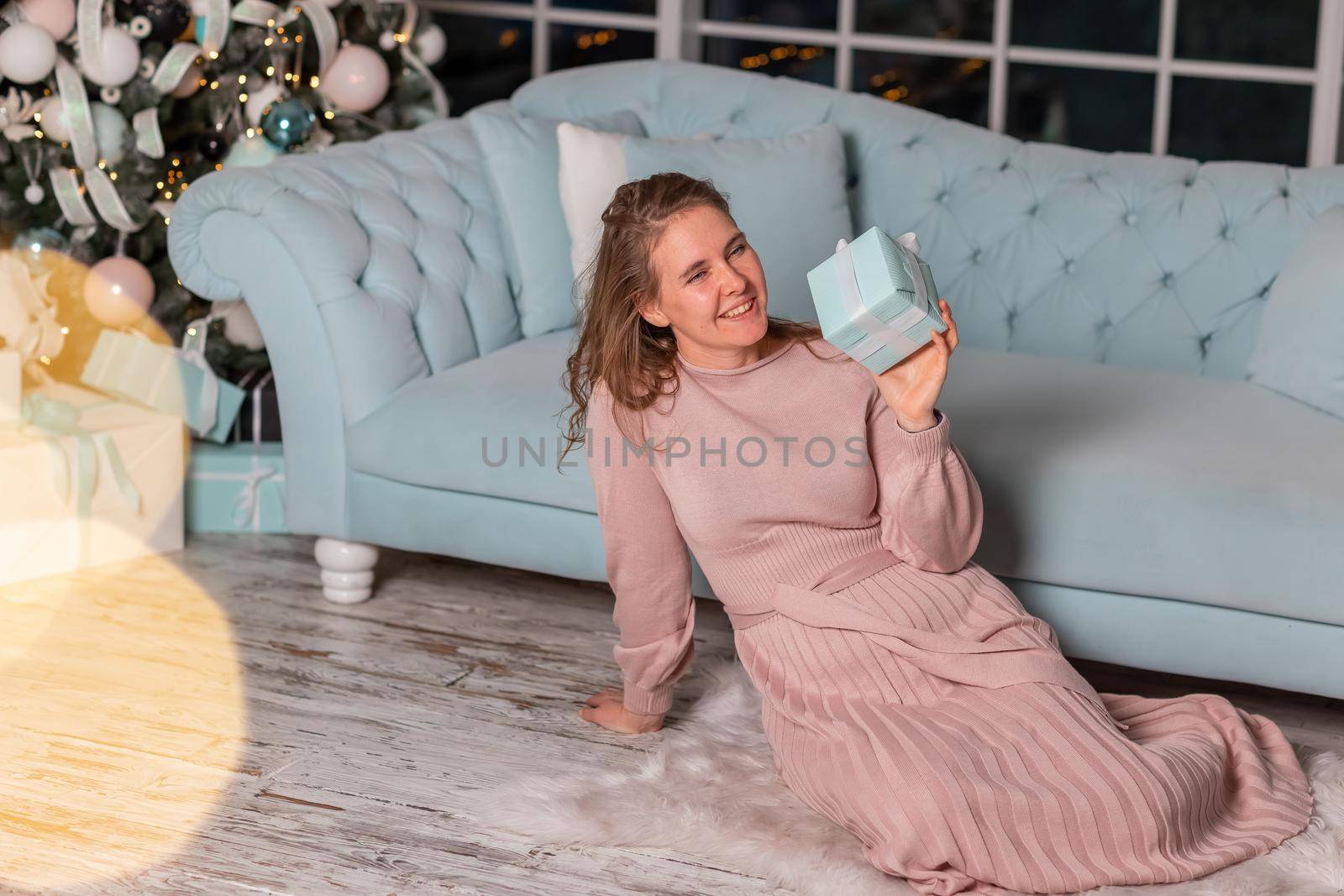Beautiful middle-aged woman opening Christmas presents near the Christmas tree on Christmas eve by Len44ik