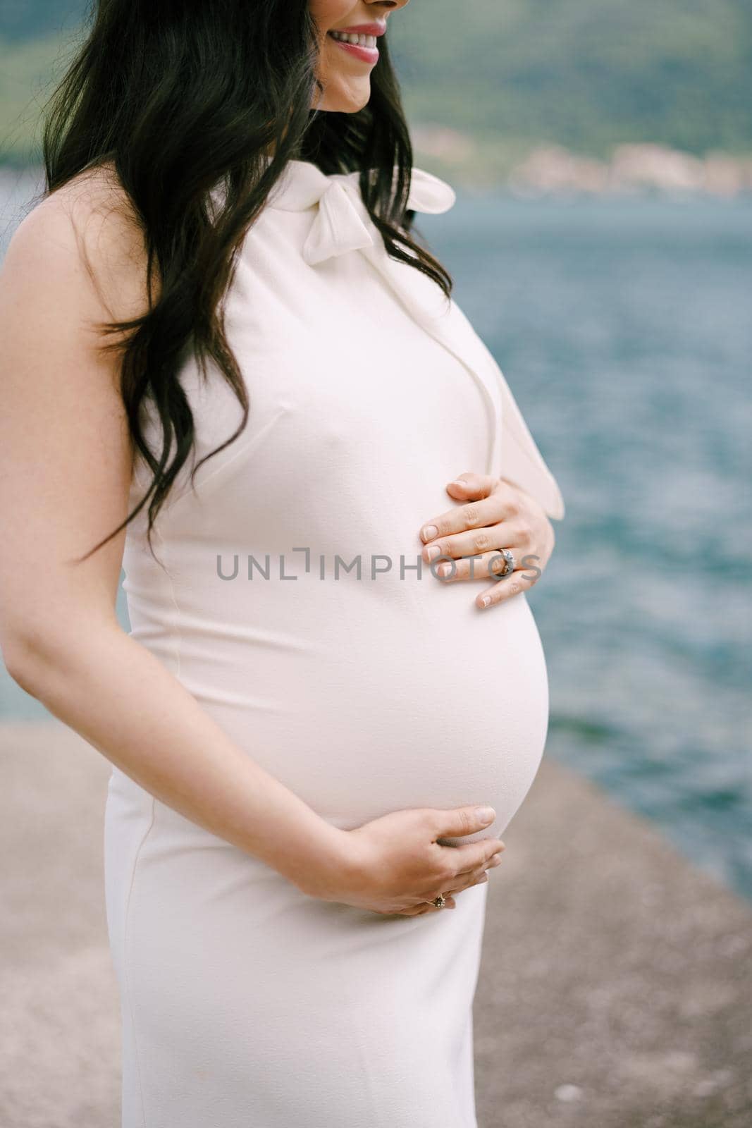 Pregnant woman in a dress stands hugging her tummy with her hands on the pier by the sea. High quality photo