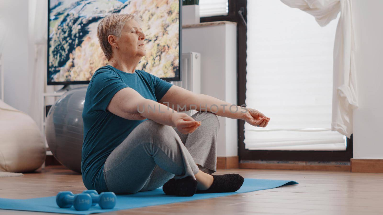 Retired woman meditating after workout training at home. Close up of old person doing meditation on yoga mat while sitting in lotus position and breathing for wellness and recreation