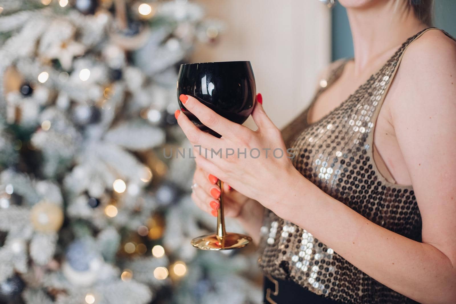 Festive female hand in bright party clothes holding goblet of alcohol beverage close-up. Attractive woman enjoying Happy New Year holiday at Christmas tree background