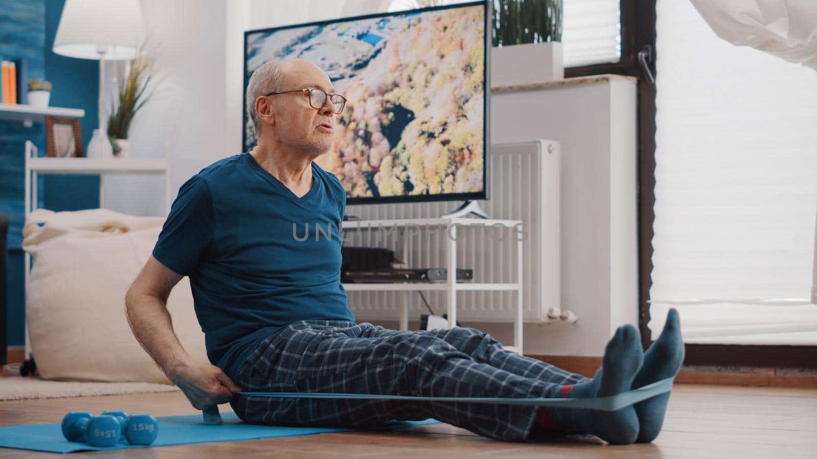 Aged man using resistance band to stretch arms and legs on yoga mat. Elder person pulling flexible elastic belt, stretching muscles and doing fitness exercise. Senior adult training