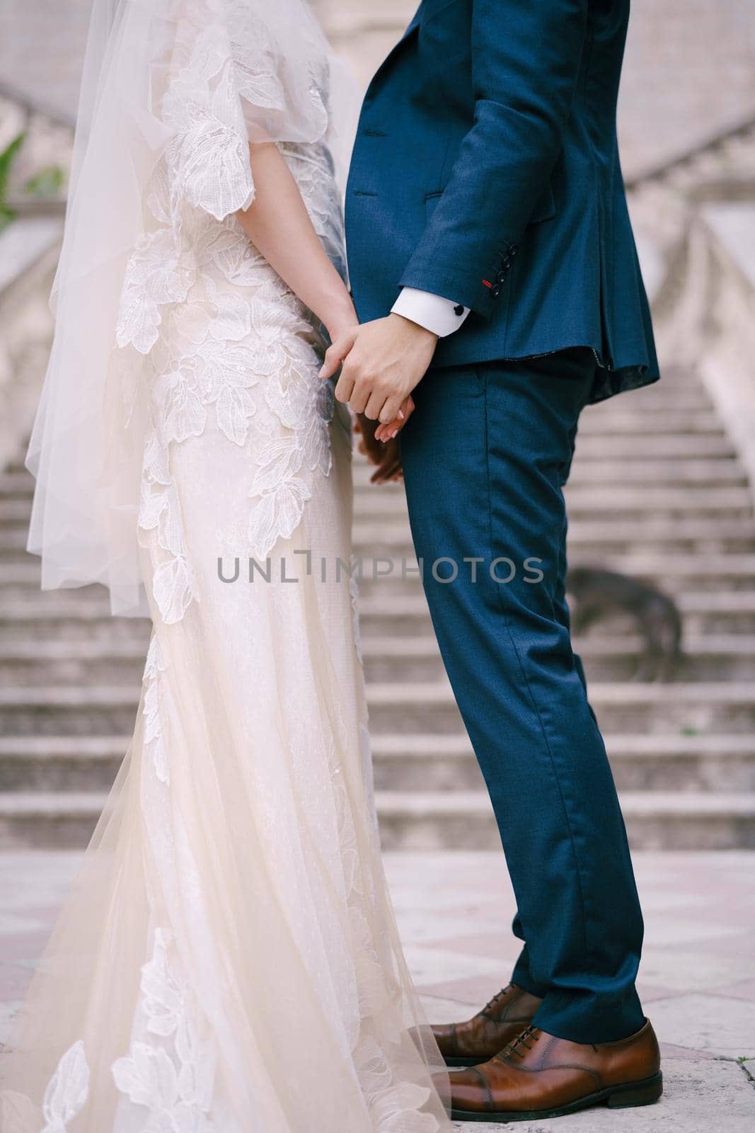 Bride and groom stand holding hands on the stone steps. Close-up by Nadtochiy