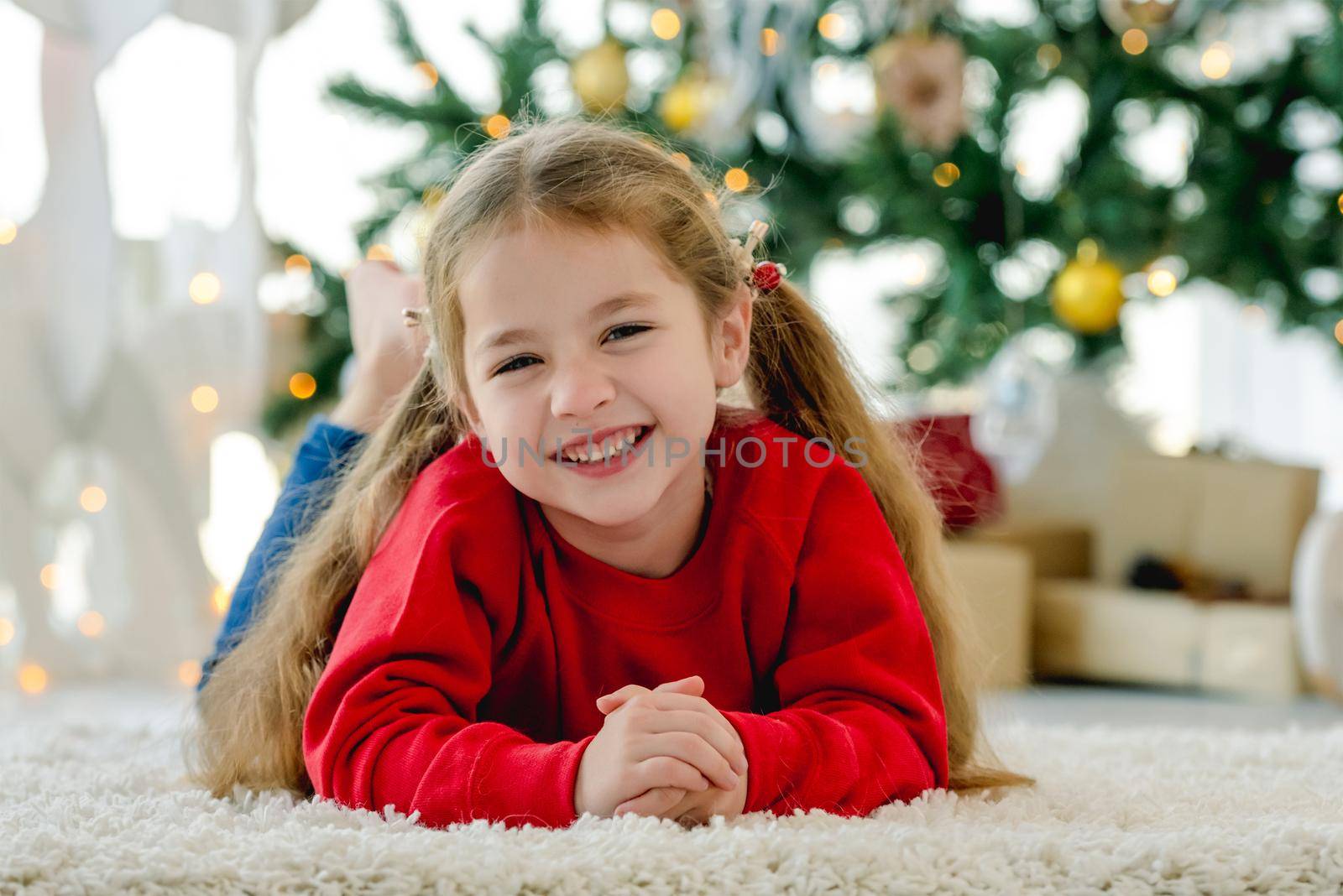 Child girl in Christmas time lying on fluffy carpet and smiling in room with decorated tree and gifts. Pretty kid at home in New Year holidays