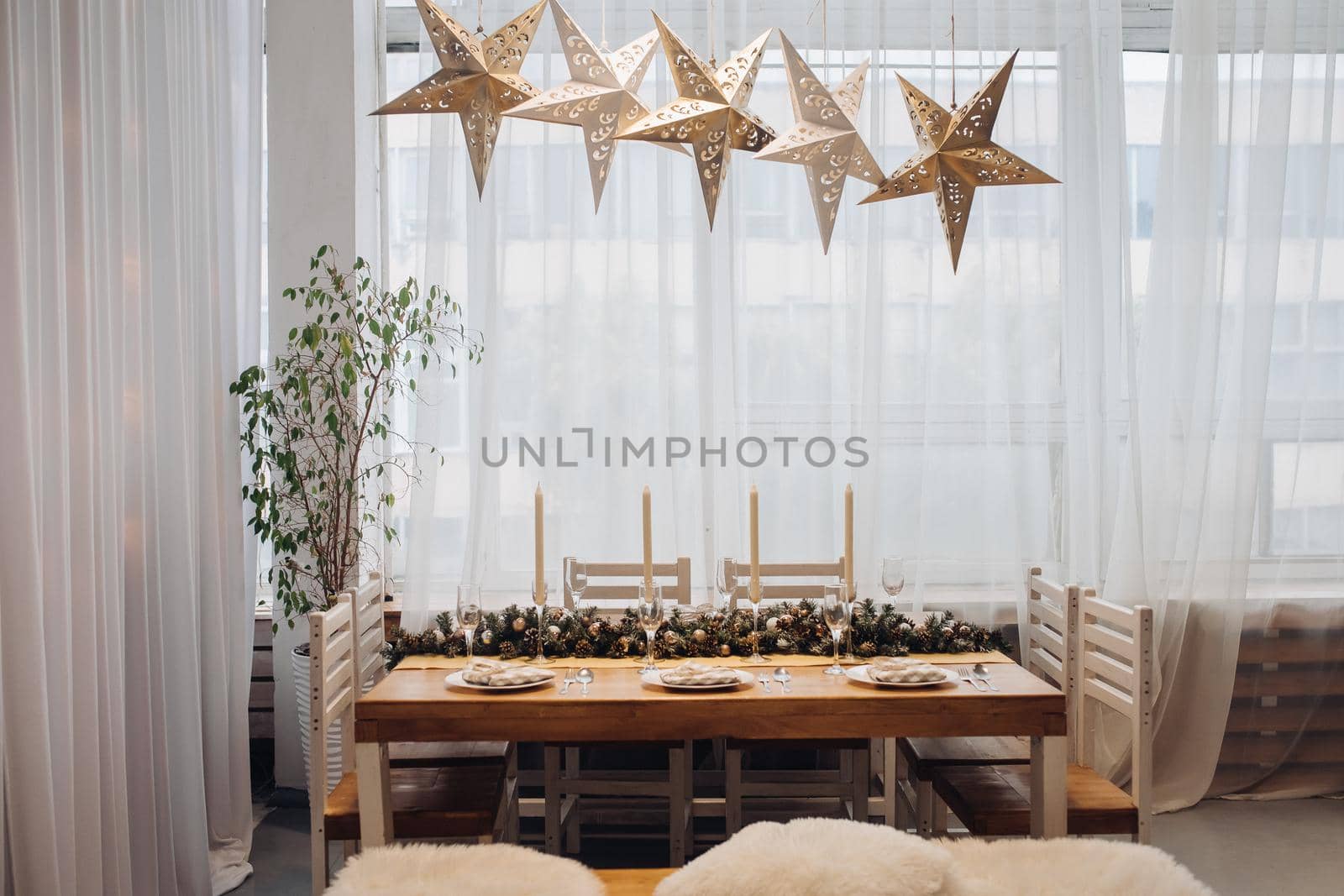View over beautifully decorated dining table for six people with fir branch with pine cones in the middle of the table, served flutes and plates. Five decorative lamps in shape of stars over the table. Christmas dinner concept.