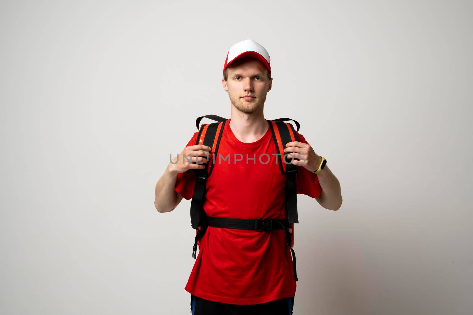 Young courier in a red uniform t-shirt and with red food thermo bag on a shoulder standing isolated on white background studio. Food delivery service. by vovsht
