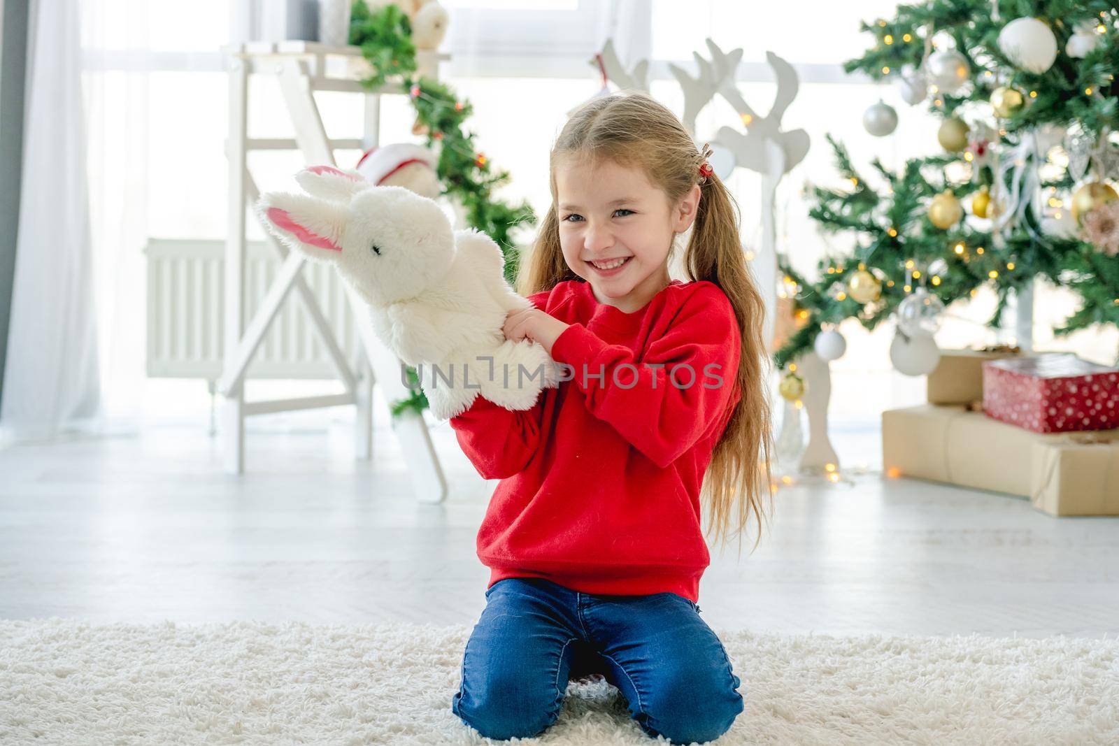 Child girl in Christmas time sitting on carpet in room with decorated tree and gifts. Pretty kid at home in New Year holidays