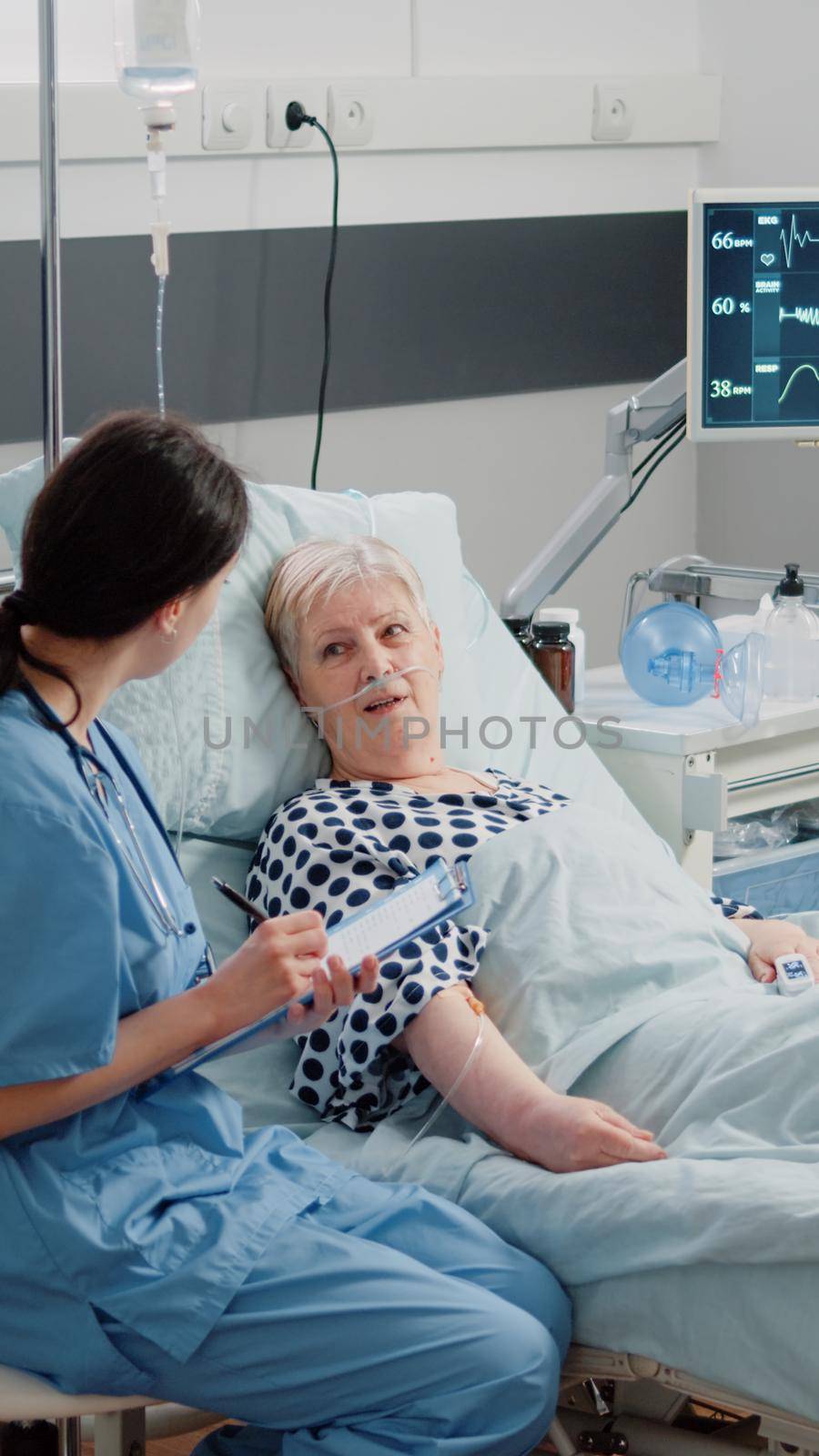 Nurse giving assistance to senior patient with disease in bed. Medical assistant and doctor doing healthcare checkup for pensioner with oxygen tube and IV drip bag in hospital ward.