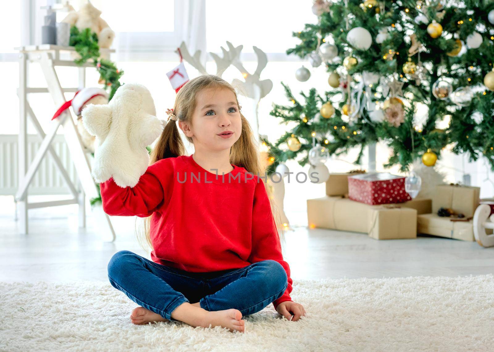 Child girl in Christmas time sitting on carpet in room with decorated tree and gifts. Pretty kid at home in New Year holidays