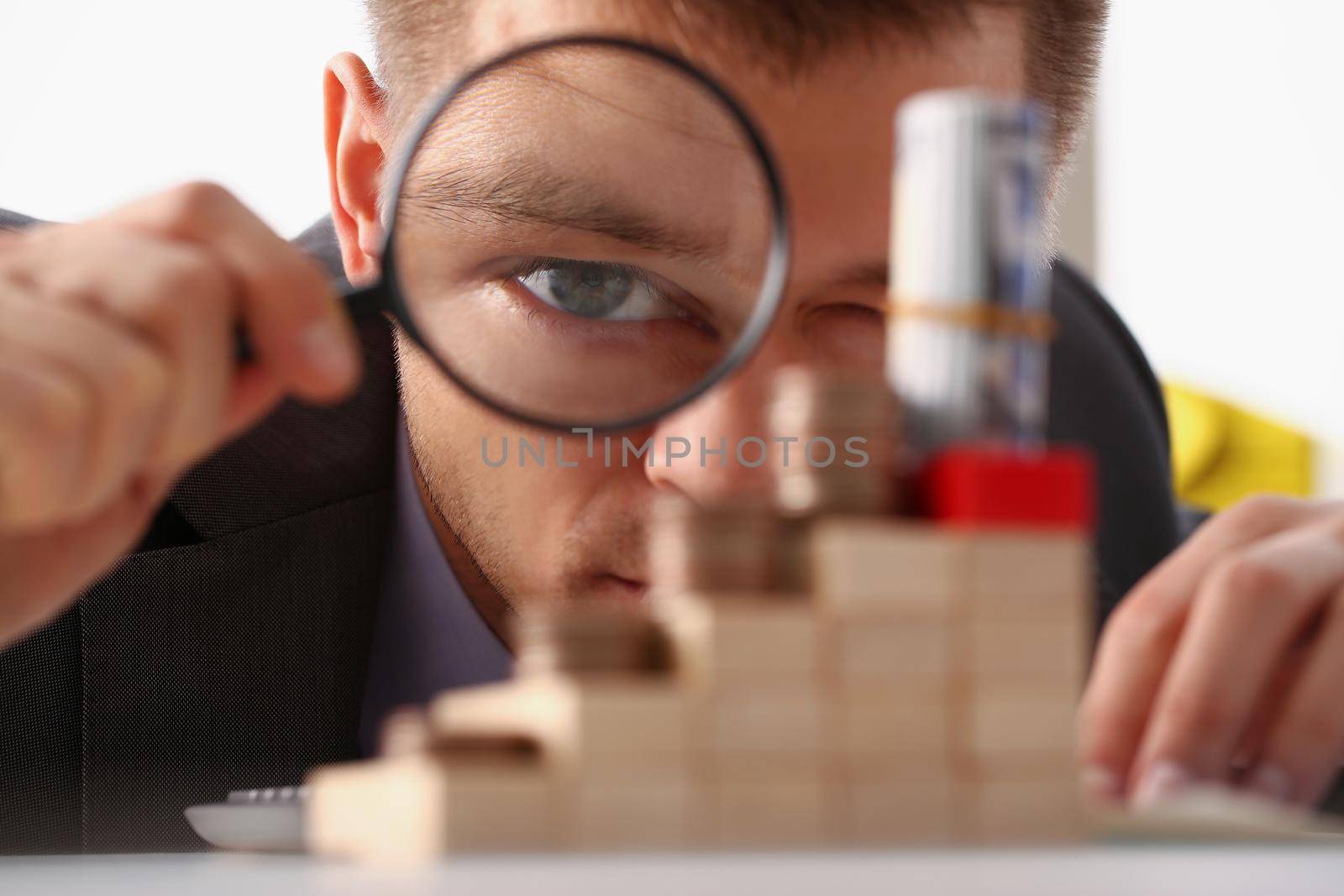 Businessman holds in hand black magnifying glass looks at tower of coins placed on wooden cubes by kuprevich