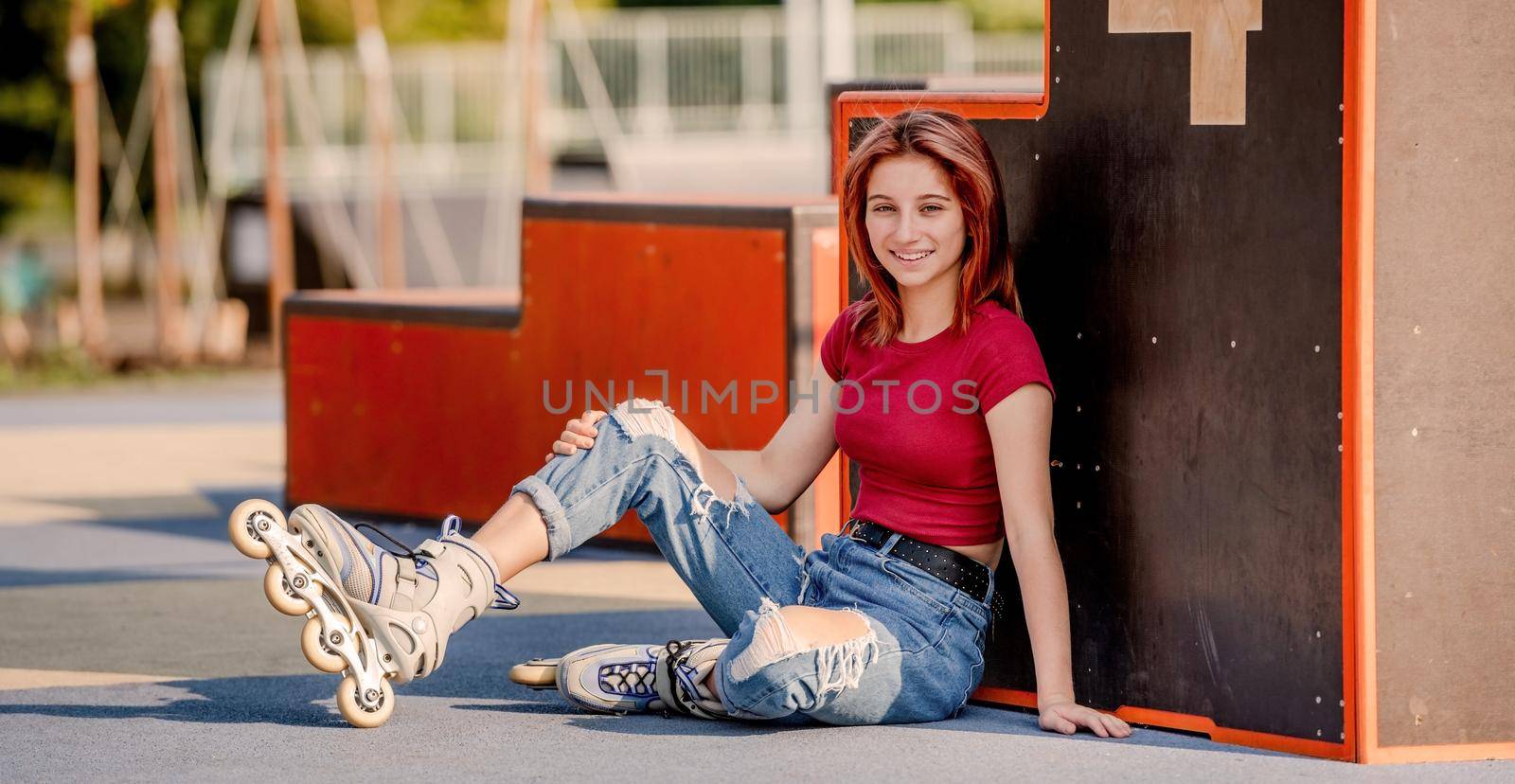 Beautiful girl wearing roller skates sitting on ground and looking at camera. Pretty female teenager skater urban portrait