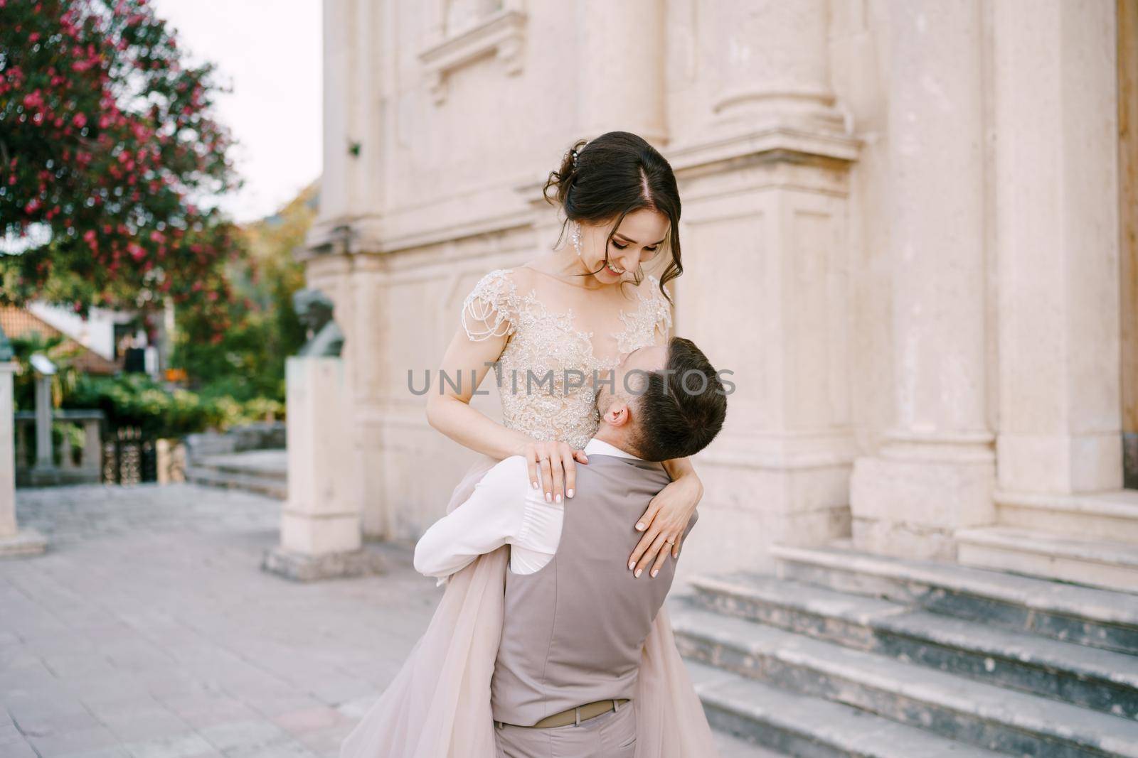 Groom holds bride in his arms in front of the marble facade of the old building by Nadtochiy