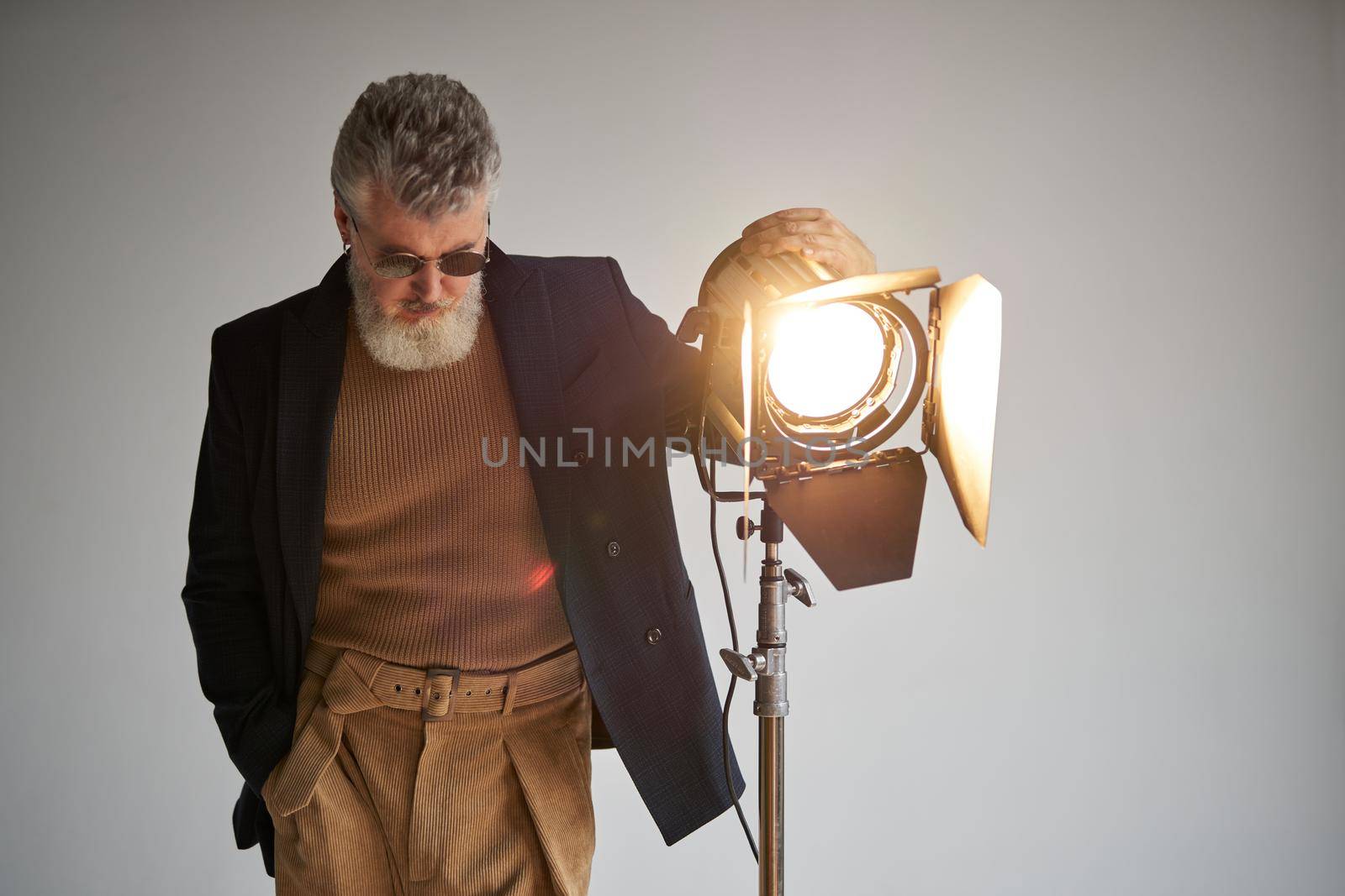 Portrait of classy bearded middle aged man dressed elegantly standing next to studio spotlight while posing for camera over white background. Fashion photoshoot, style concept