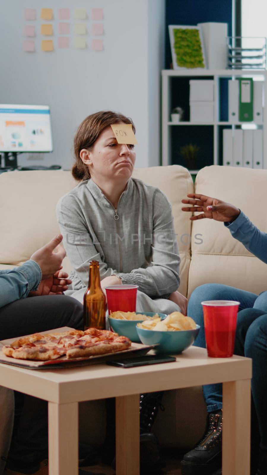 Cheerful coworkers playing guessing game with sticky notes on forehead after work for entertainment. Workmates enjoying fun activity with beer drinks, snacks and play after hours.