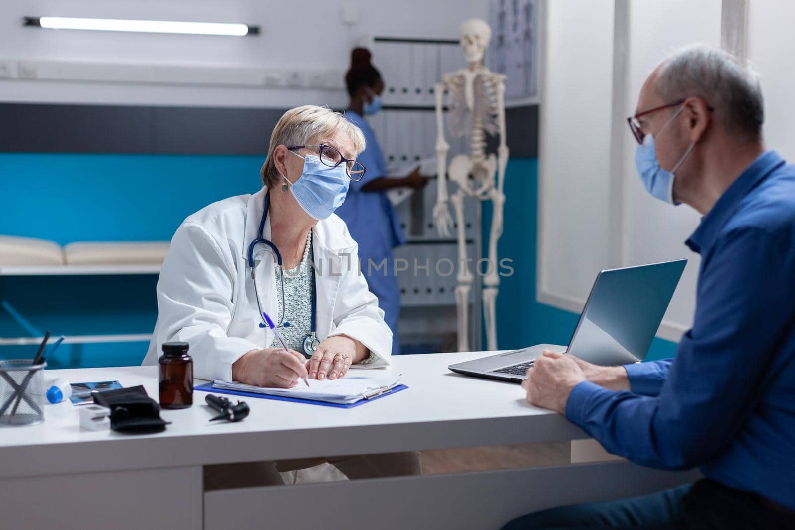 Woman doctor with face mask taking notes on papers at medical consultation with man by DCStudio