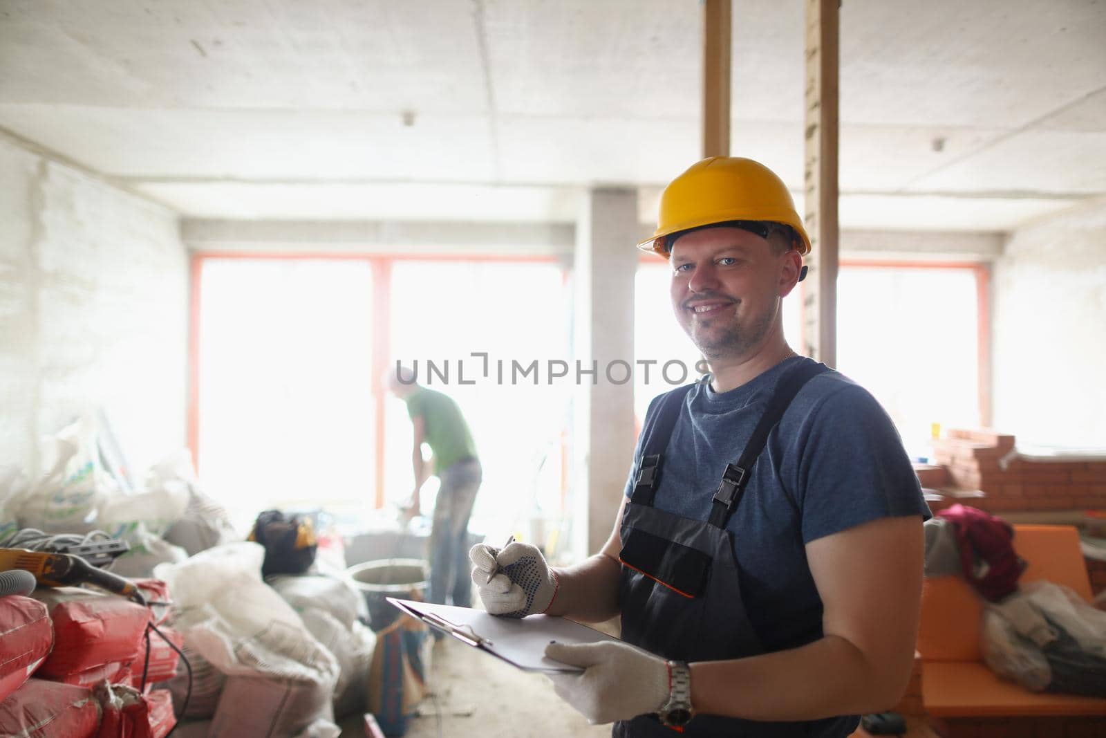 Portrait of smiling professional worker hold clipboard and list work materials. Joyful worker wear uniform and protective helmet. Construction site concept