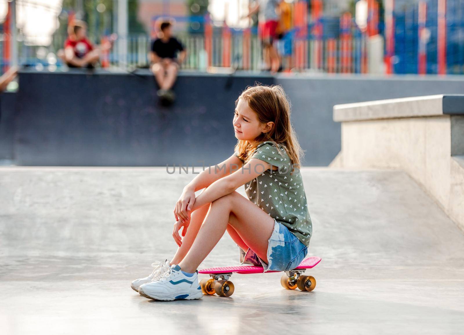 Preteen girl sitting on skateboard outdoors and looking back. Female skater child close to city riding ramp