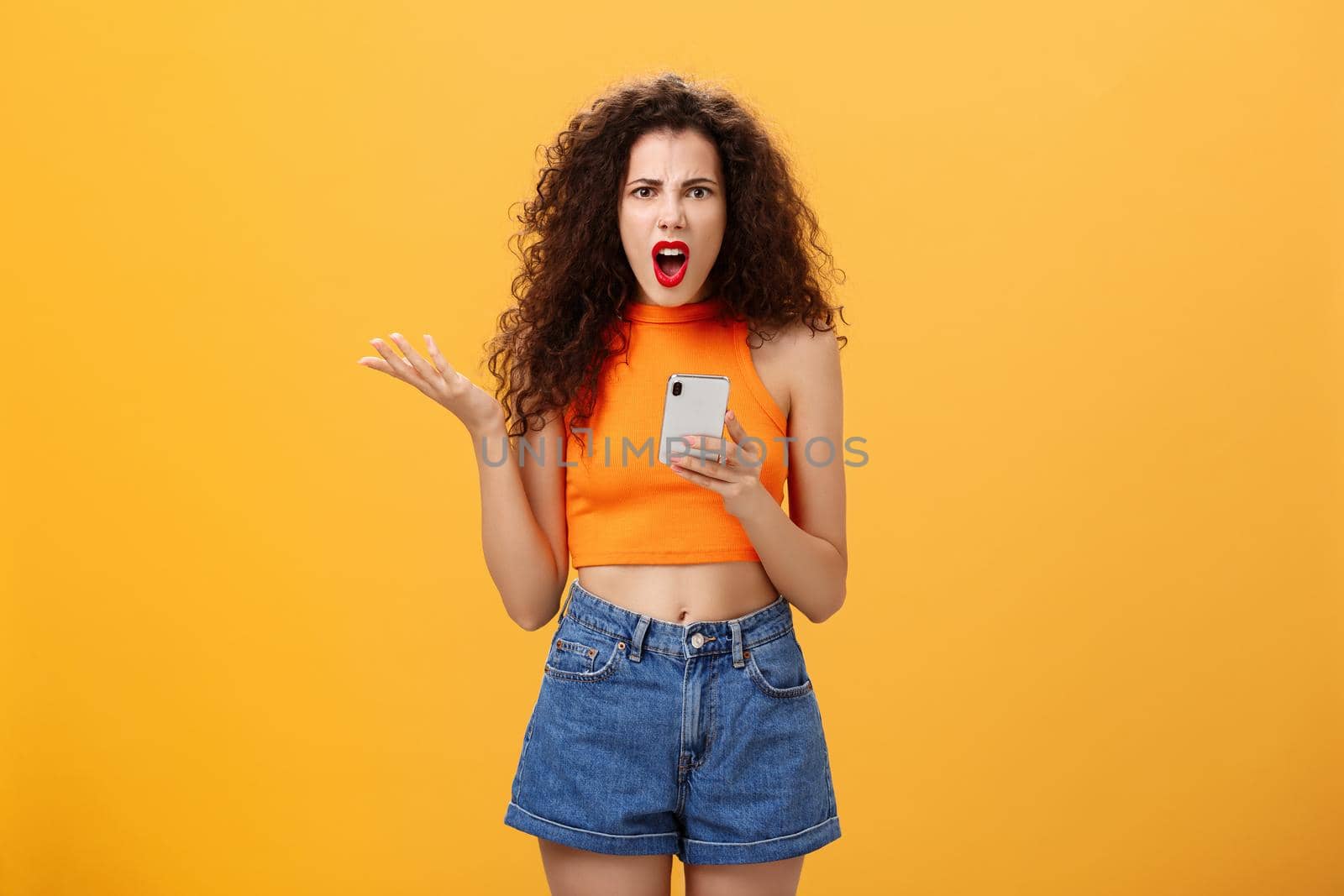 Portrait of displeased pissed and irritated caucasian female with curly hairstyle in red lipstick and orange cropped top holding smartphone and gesturing with palm annoyed complaining on dumb message by Benzoix