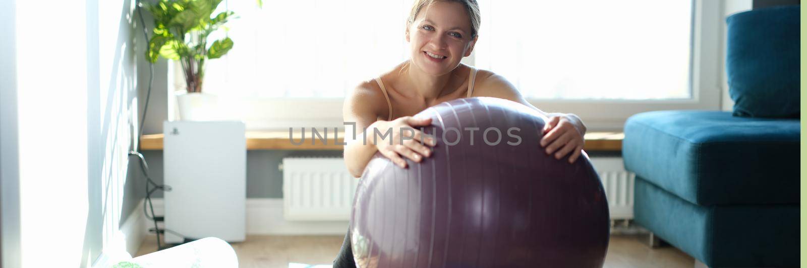 Portrait of happy young woman with large gymnastic ball on rug at home. Fitball exercises at home concept