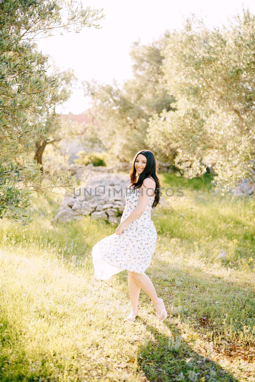 Smiling pregnant woman standing on green grass holding the hem of her dress. High quality photo