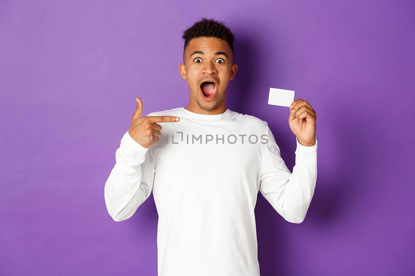 Portrait of amazed african-american man pointing at credit card, recommend bank loan, standing over purple background.