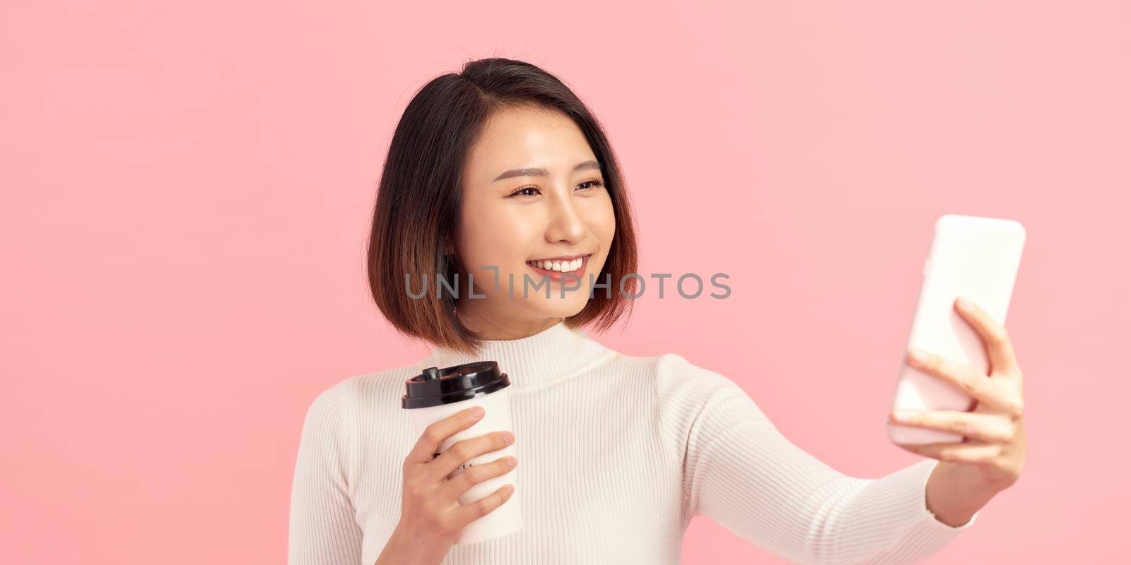 Portrait of a smiling attractive woman taking a selfie while holding take away coffee cup over pink background