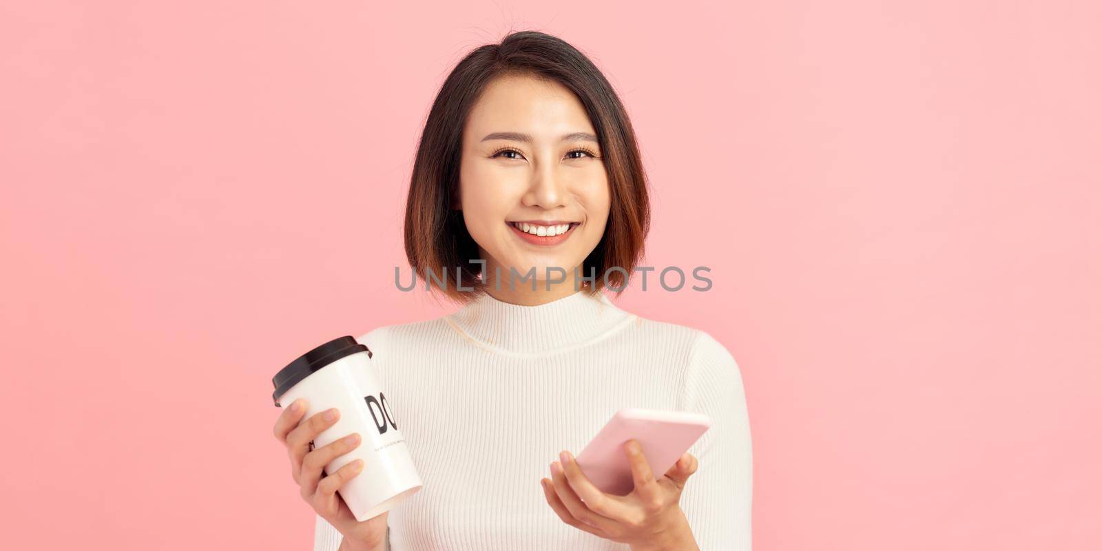 Young asian woman over isolated pink background holding coffee to take away and a mobile by makidotvn