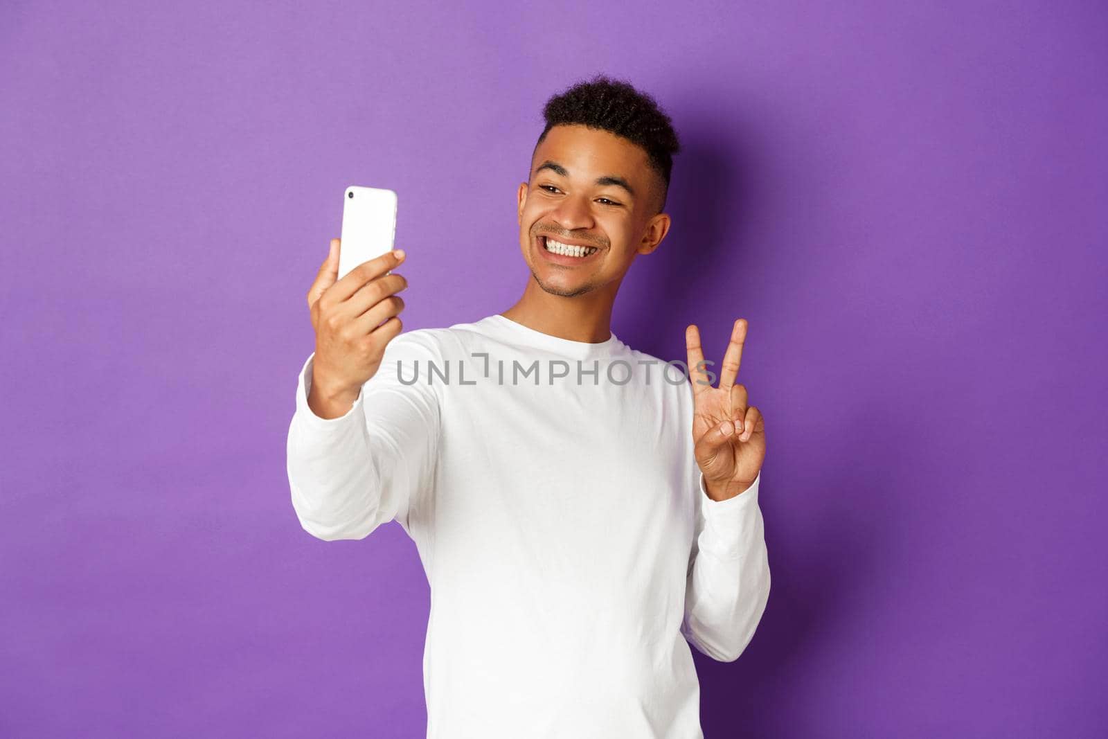 Cheerful african-american male blogger, taking selfie on smartphone, showing peace sign and smiling at mobile phone camera, standing over purple background.
