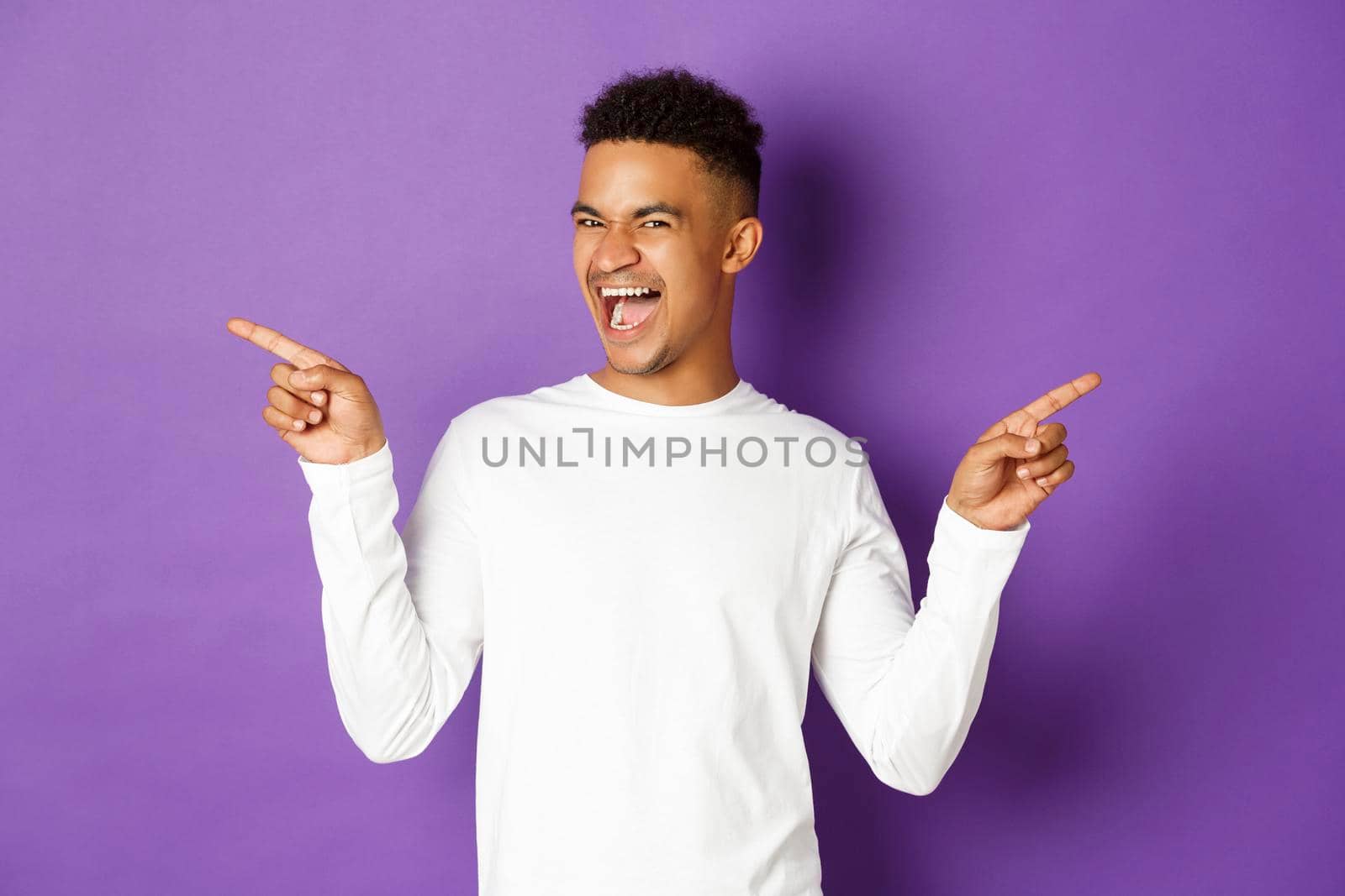 Image of cheerful african-american guy in white sweatshirt, showing two variants, pointing fingers sideways at copy space and smiling, demonstrate products, purple background.