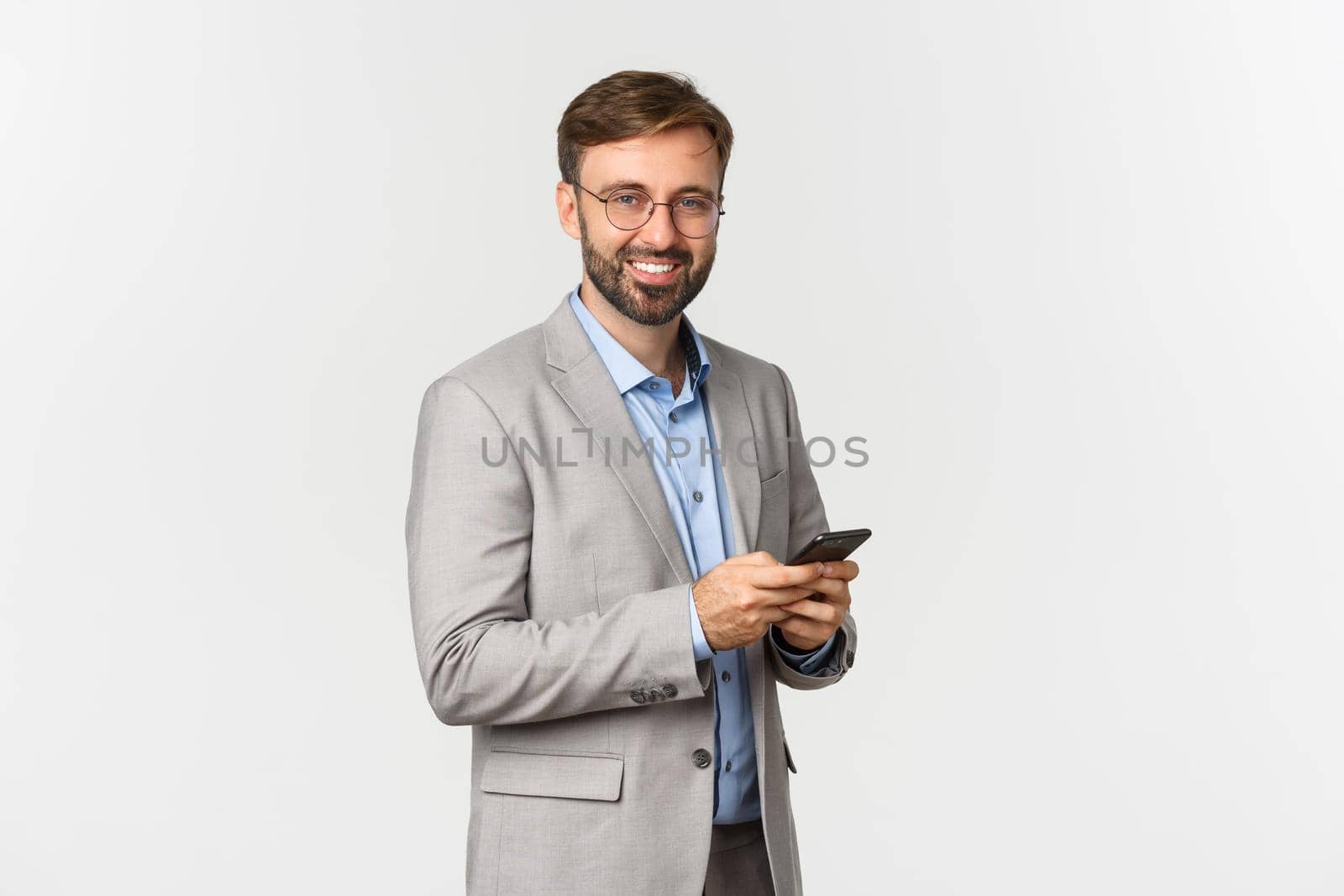 Handsome businessman with beard, wearing glasses and suit, holding mobile phone and smiling at camera, standing over white background by Benzoix