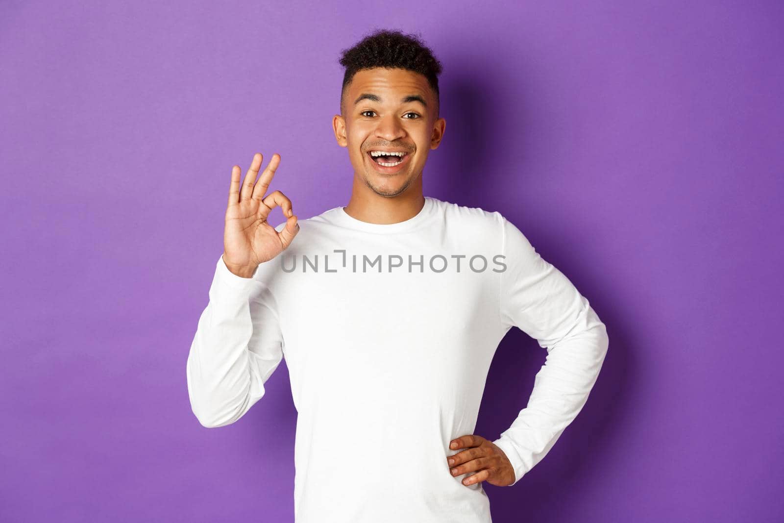Portrait of satisfied smiling african-american man, wearing white sweatshirt, showing okay sign in approval, like and agree with something, standing over purple background.