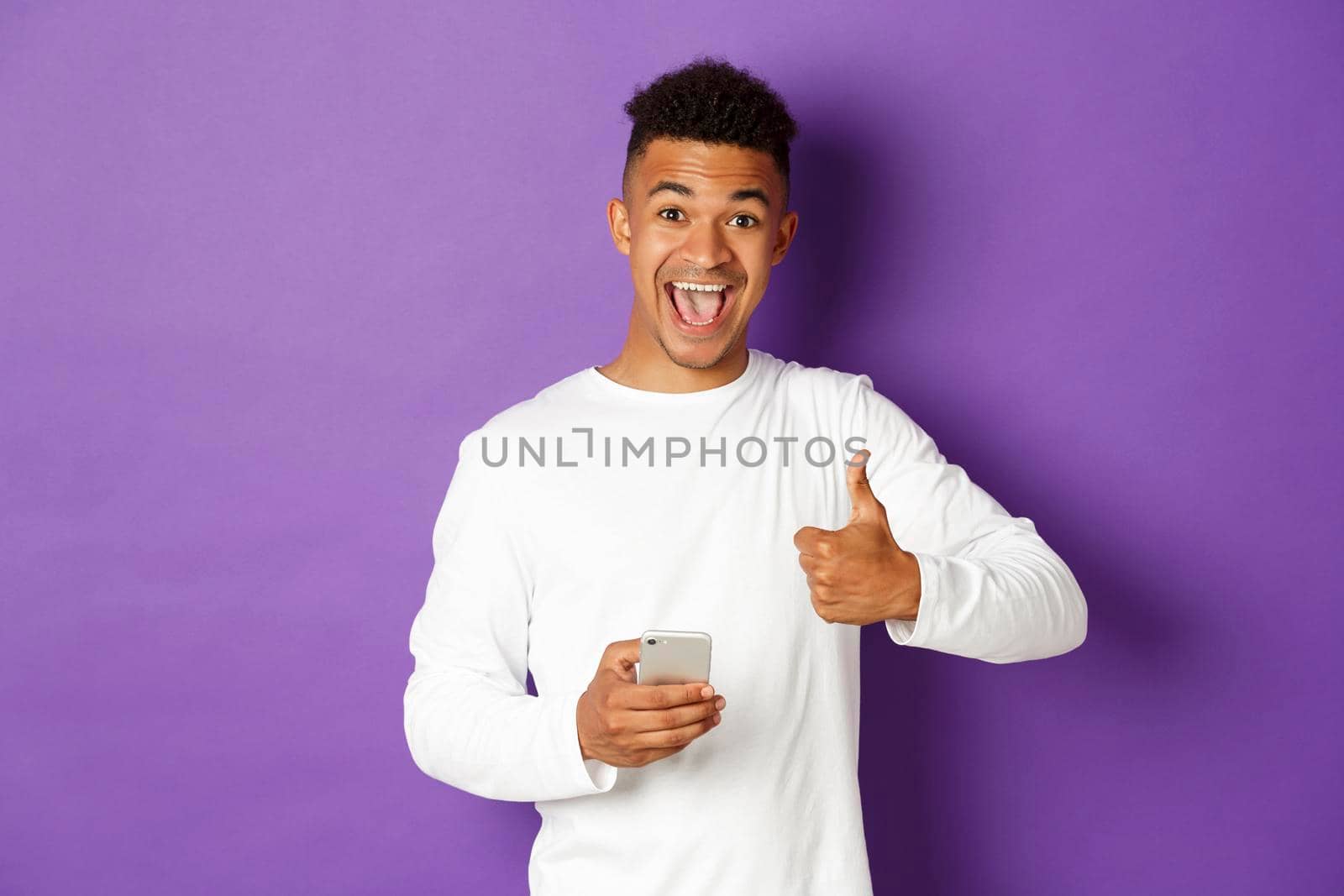 Image of amazed and satisfied african-american guy, holding mobile phone and showing thumbs-up in approval, like something, standing over purple background.